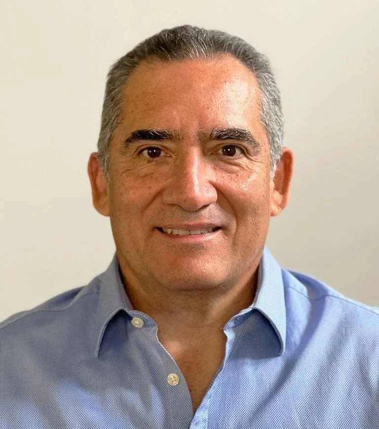 Portrait of a middle-aged man with short, gray hair, wearing a light blue button-up shirt, smiling, against a plain light-colored background.