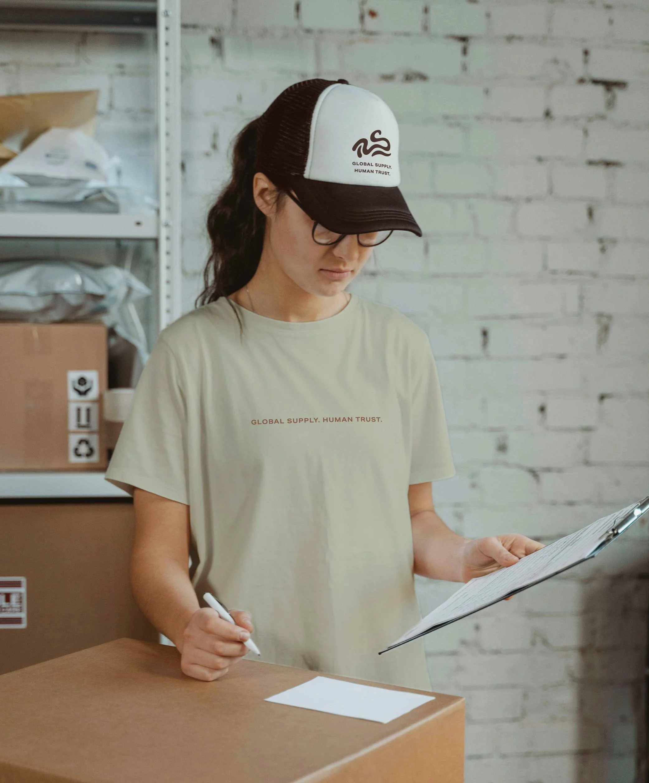 A woman with dark hair, glasses, and a white and black cap reading a document. She is wearing a beige T-shirt with brown text.