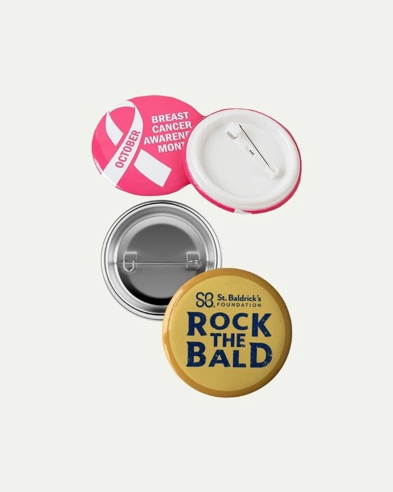 Three round buttons with different designs and messages: one pink with a white ribbon and text about Breast Cancer Awareness Month, one with a metal back and pin, and one yellow with blue text saying 'Rock the Bald' from St. Baldrick's Foundation.