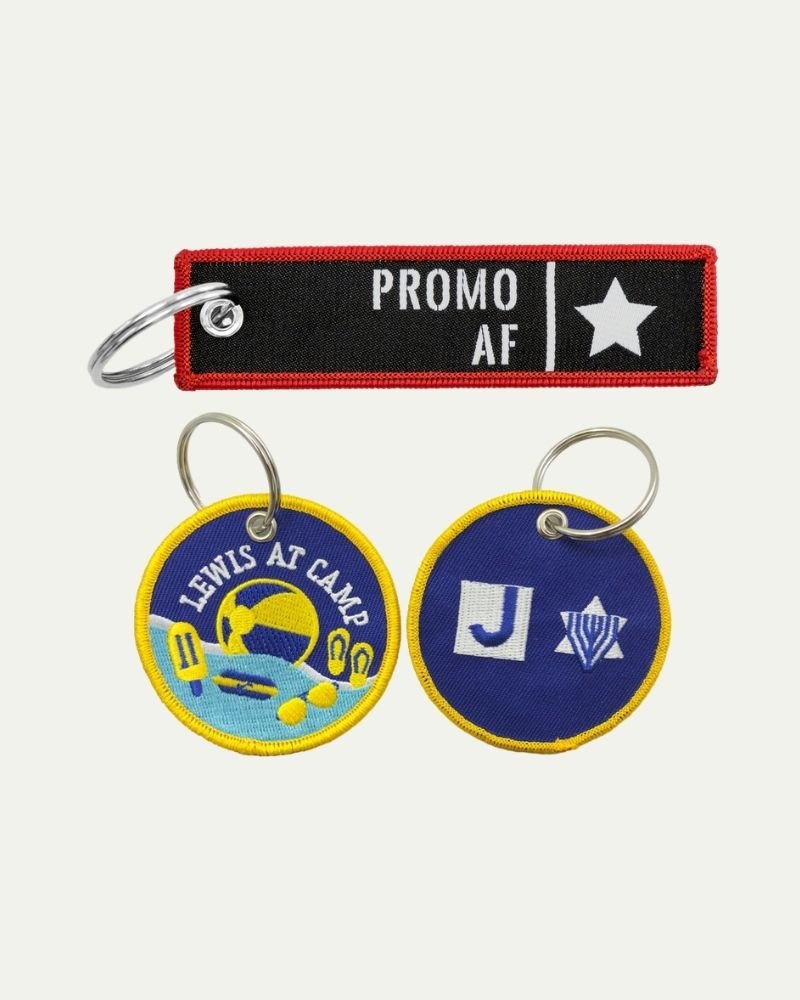 Three embroidered keychains: a black and red one reading 'PROMO AF' with a star, and two with yellow borders; one showing 'LEWIS AT CAMP' with a basketball and volleyball, the other with a 'J' and a V-shaped symbol.