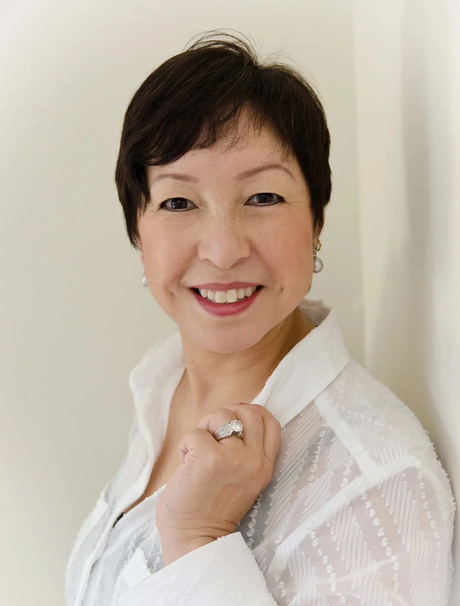 Close-up of a smiling woman with short dark hair wearing a white textured blouse and pearl earrings, holding her hand near her chin with an engagement ring visible.