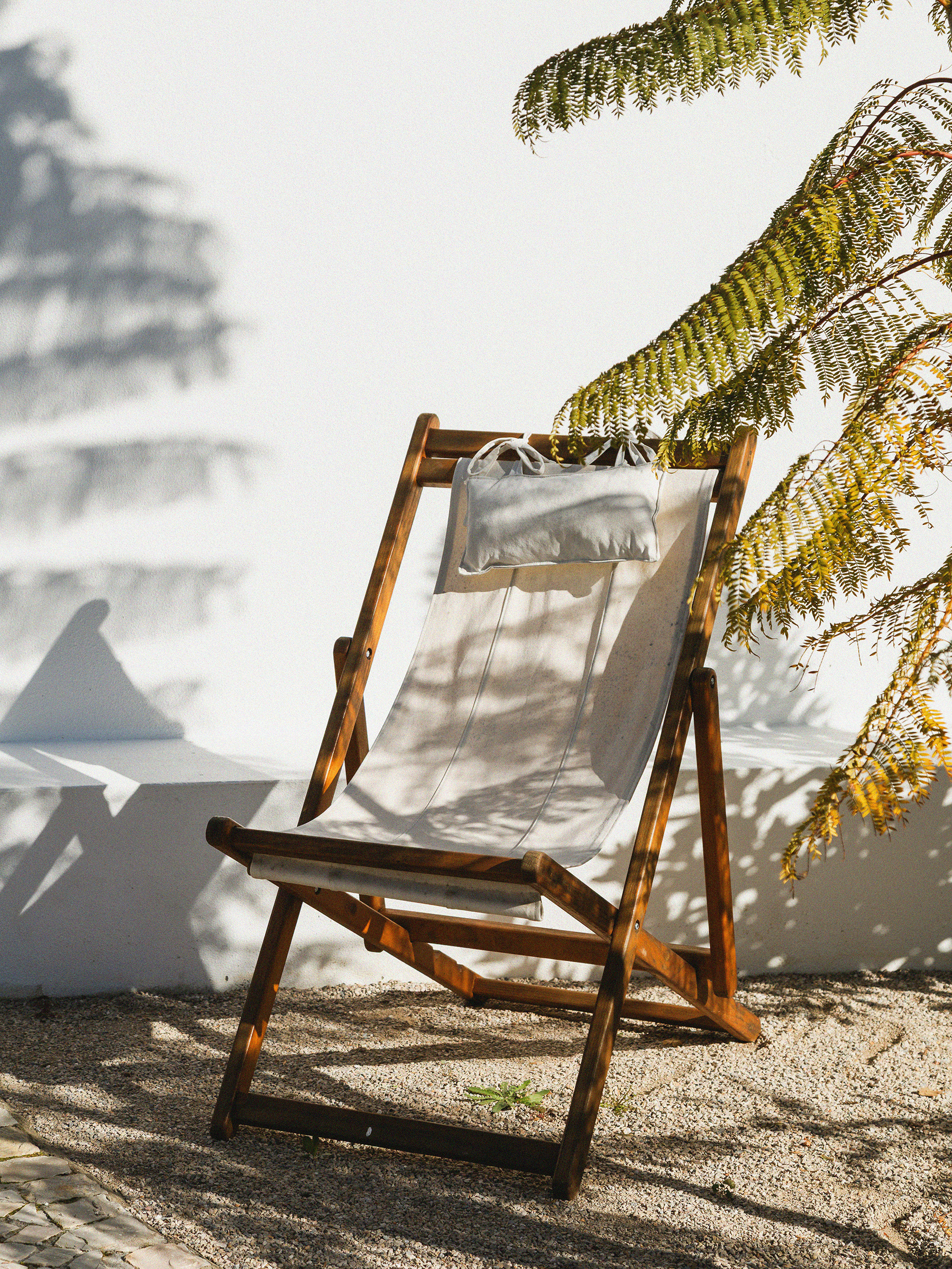 A foldable wooden chair with a white cushion and pillow, positioned on sandy ground, with shadows of fern leaves cast on a white wall nearby.