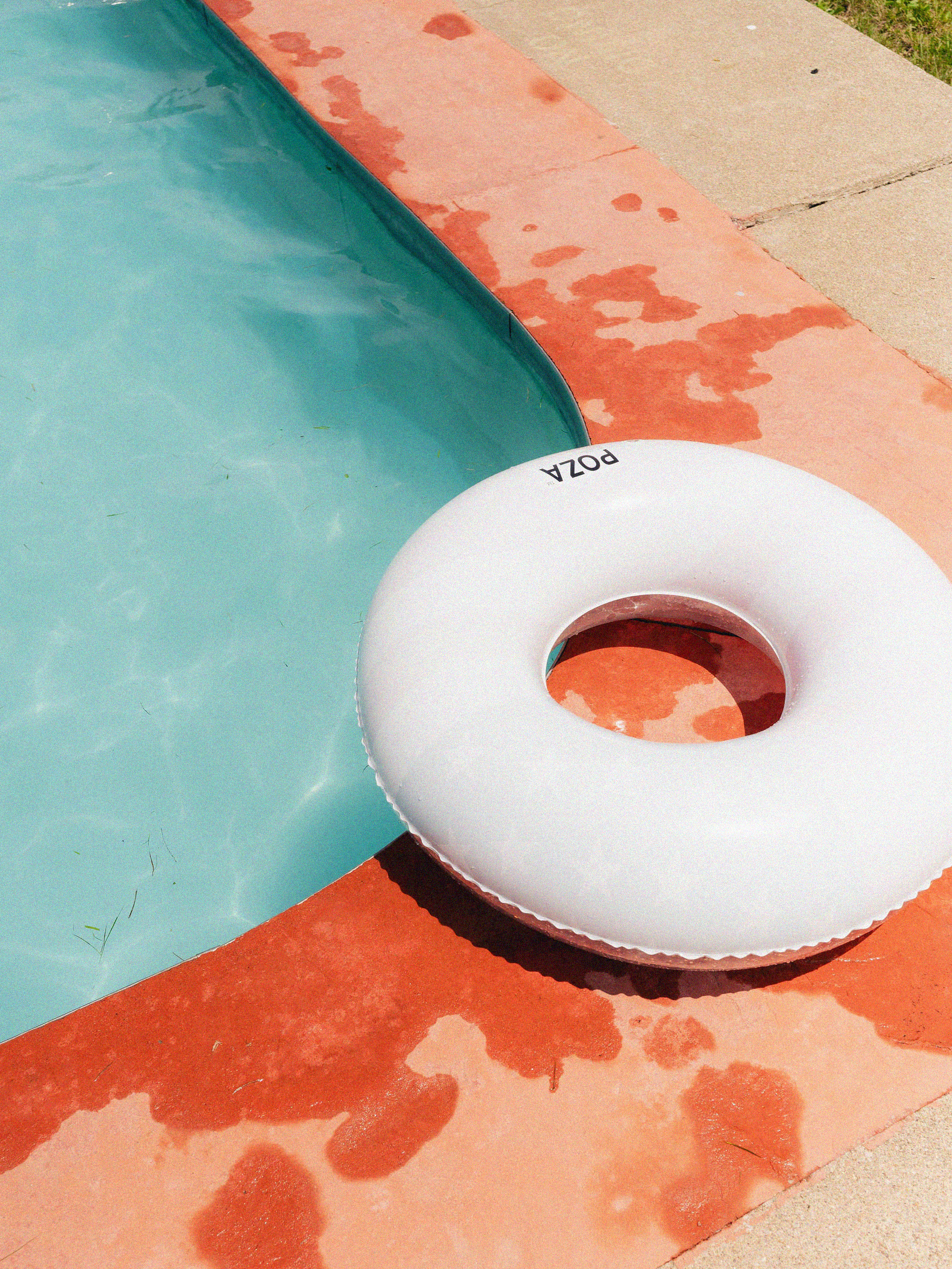 A white inflatable pool ring with the word 'POZA' on it rests on the edge of a swimming pool with clear water. The pool is surrounded by a red and concrete pool deck, with evidence of water splashes and wet patches on the surface.