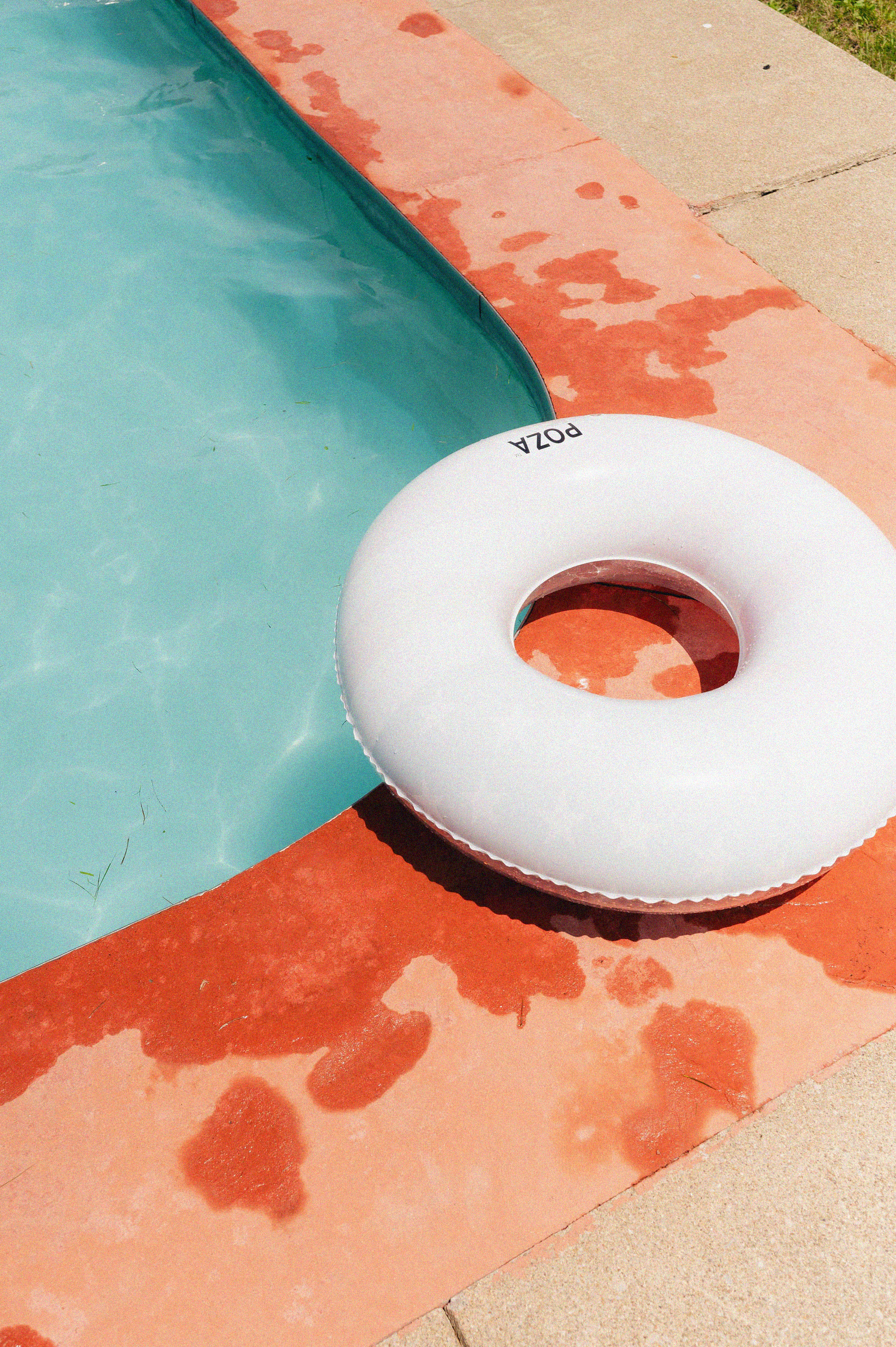 An inflatable white pool float with a hole in the center resting on a red colored pool deck next to a swimming pool filled with water.