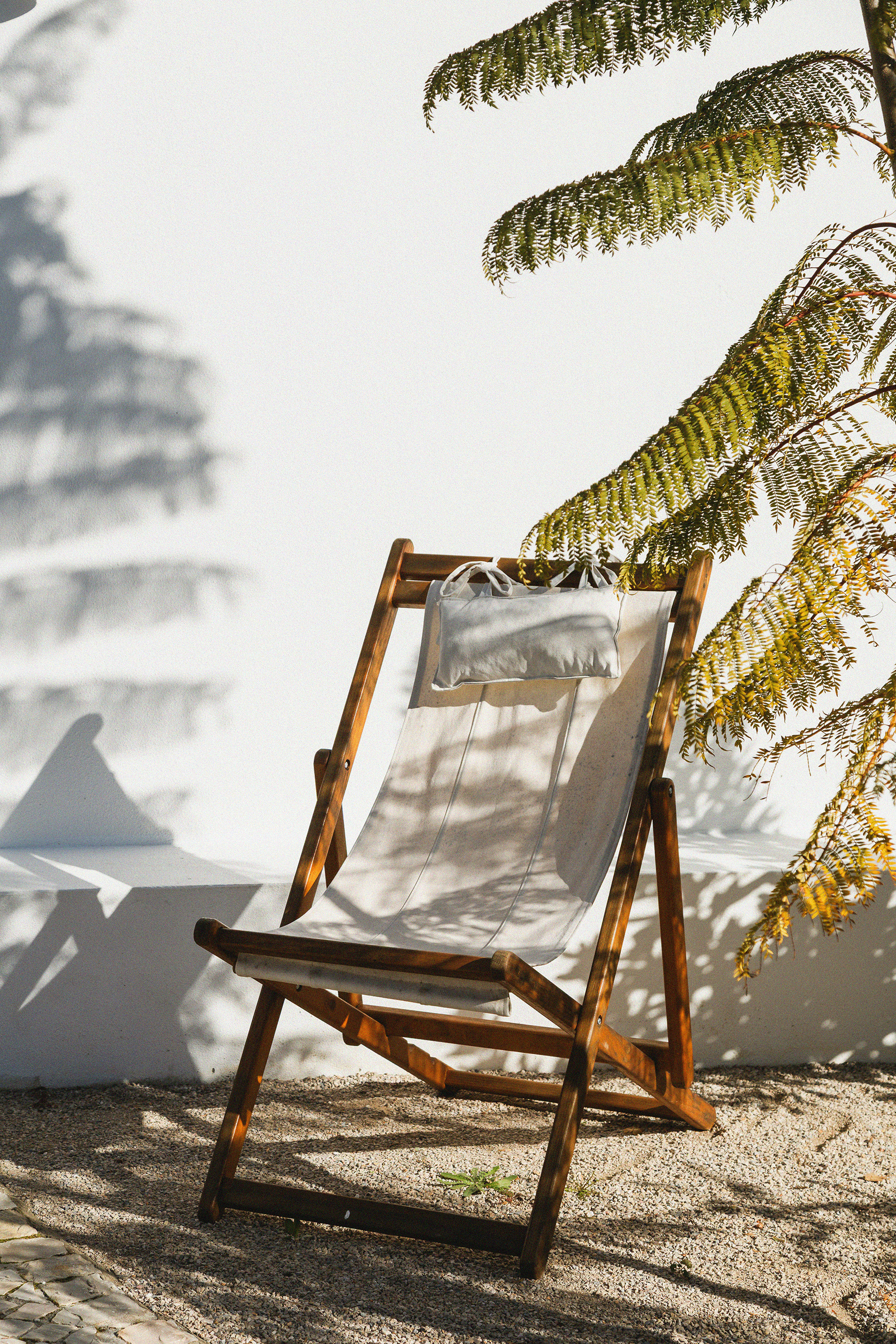 A wooden deck chair with a white pillow, set outdoors on a sandy surface, casting shadows from nearby fern leaves.