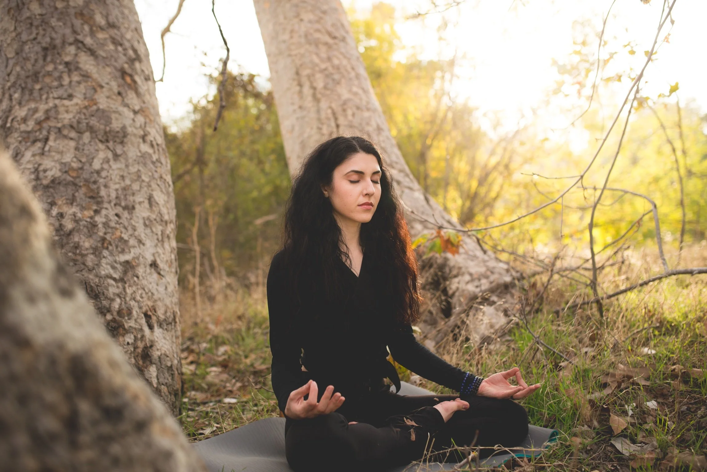 A young woman with long dark hair practicing yoga outdoors in a forest, sitting cross-legged on a mat with her eyes closed in a meditative pose during sunset.