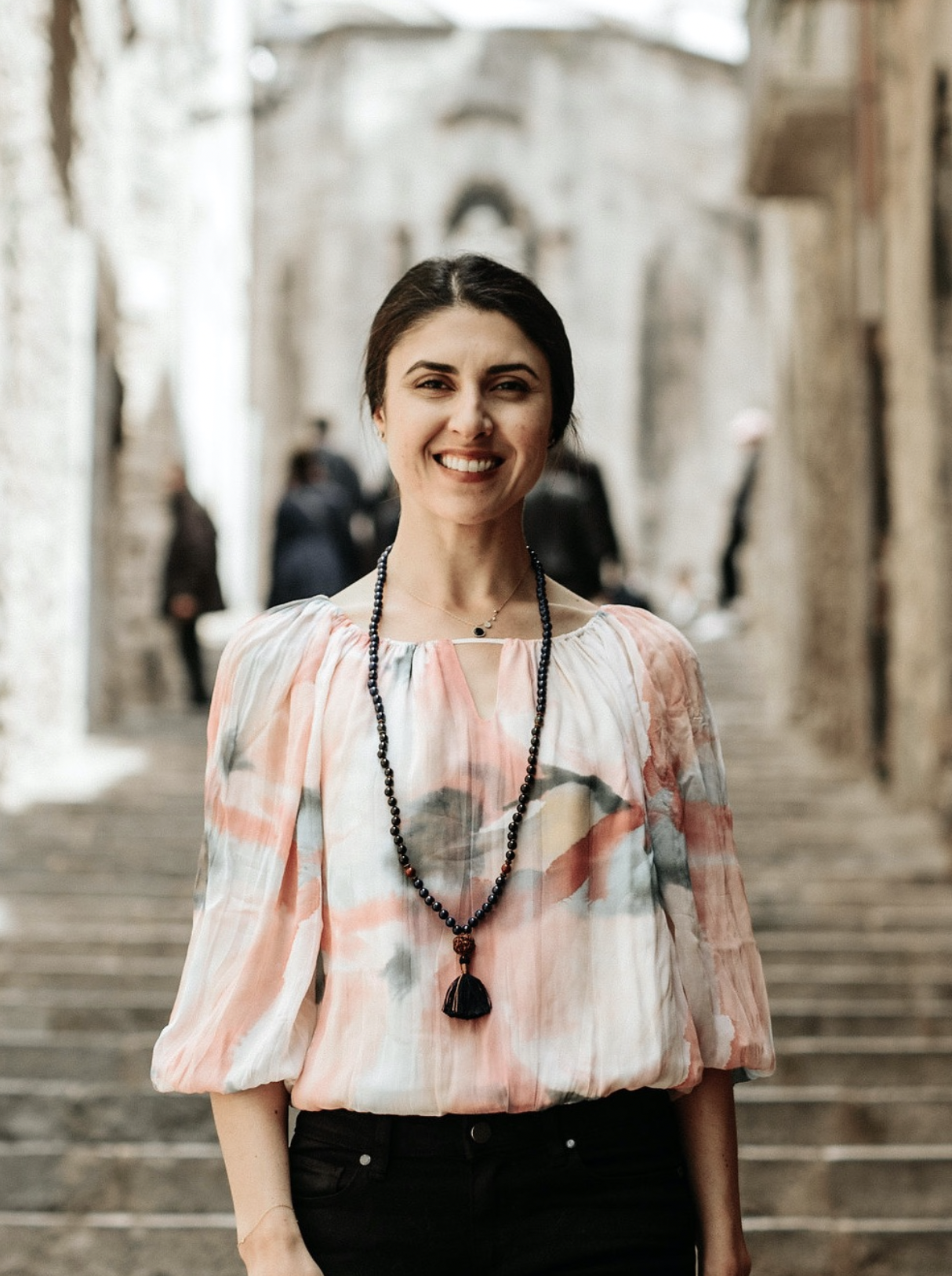 A smiling woman with dark hair tied back, wearing a light-colored, patterned blouse and a long beaded necklace, standing outdoors on stone stairs with blurred people and historic buildings in the background.