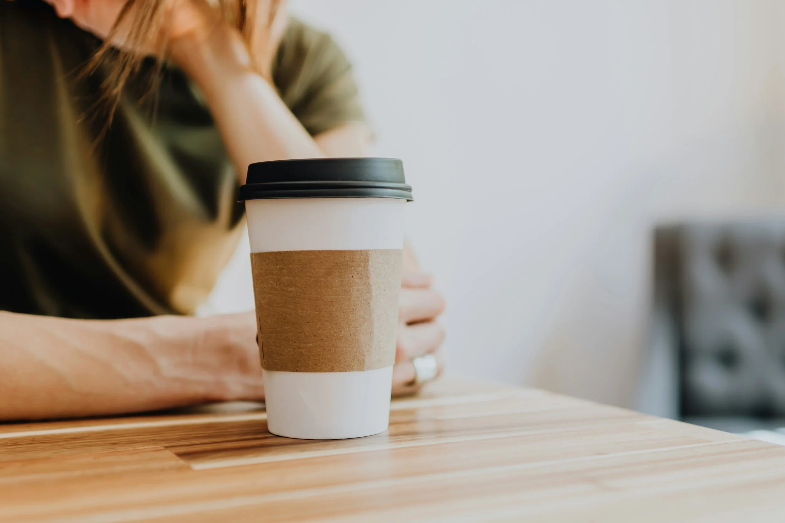 A person resting their head on one hand at a wooden table with a to-go coffee cup in the foreground.