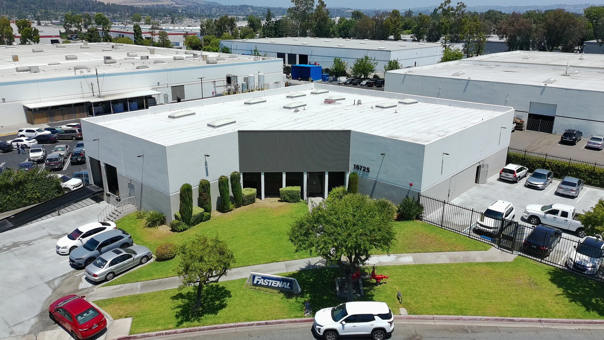 An aerial view of a commercial building with a white roof, surrounded by parked cars and green landscaping, with additional industrial buildings and trees in the background.
