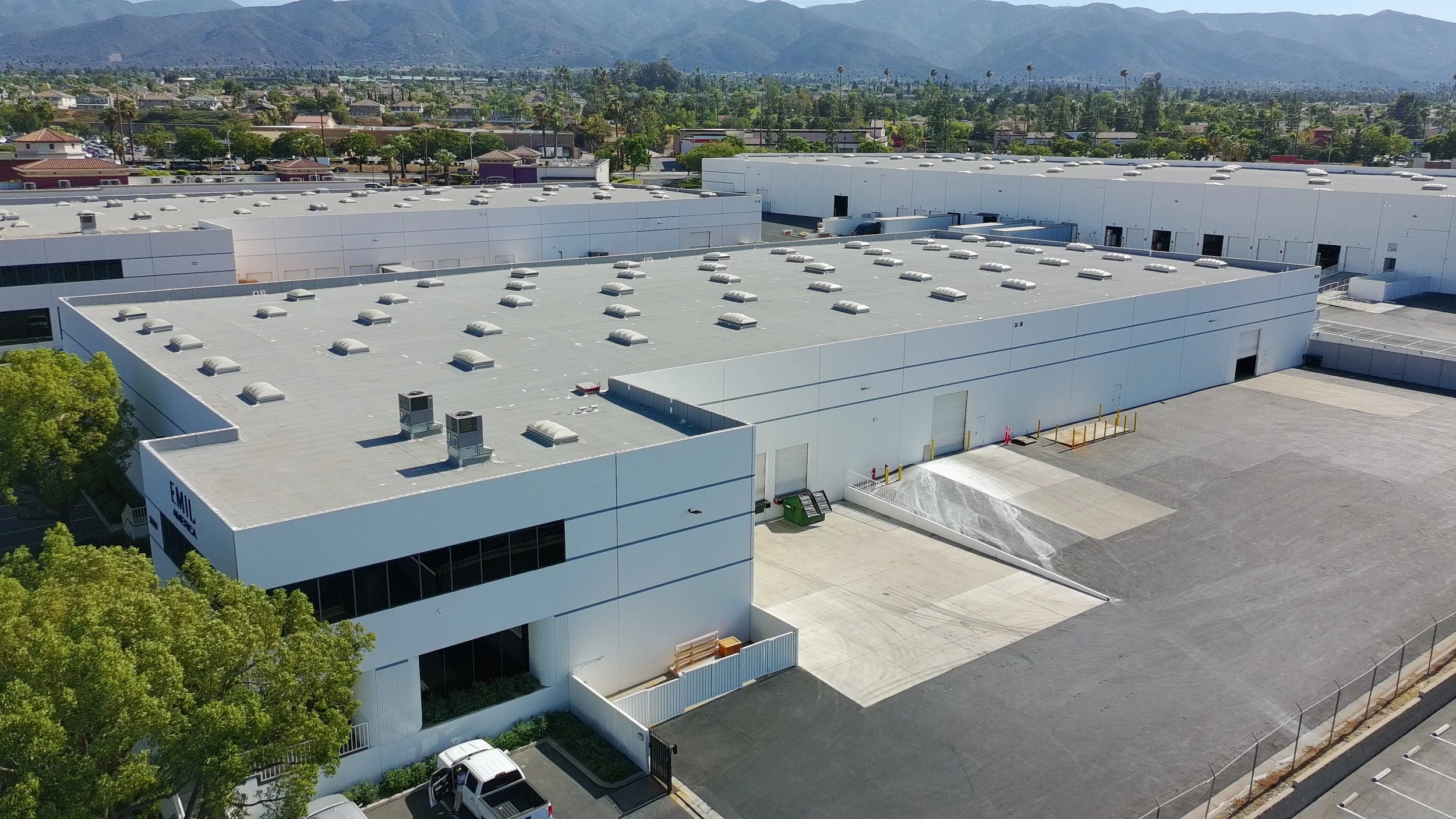 Aerial view of a large white industrial building with a flat roof, numerous skylights and ventilation units, surrounded by parking areas, trees, and a cityscape with mountains in the background.