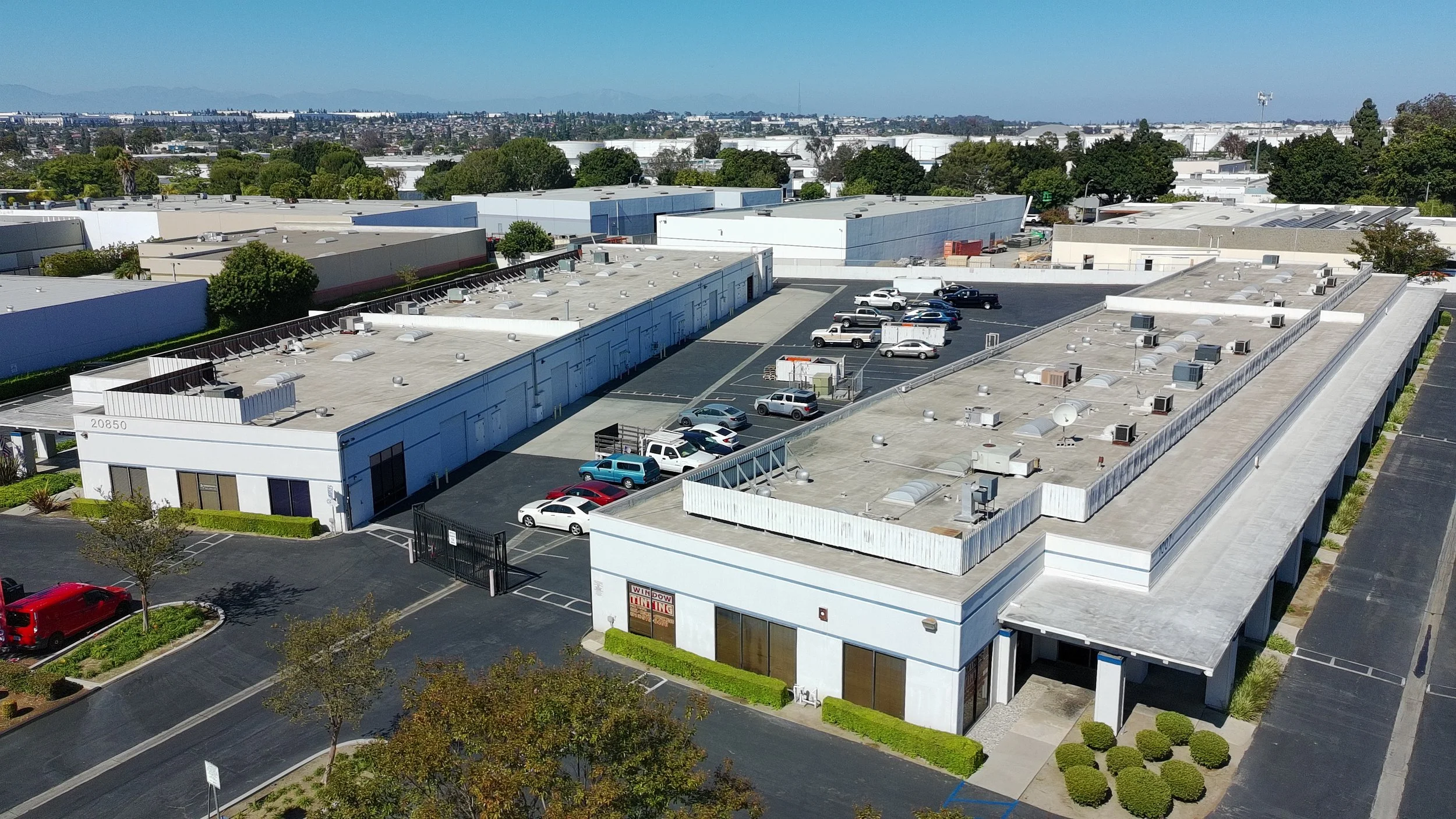 Aerial view of a commercial warehouse complex with multiple flat-roofed buildings, parking lot, and surrounding greenery.