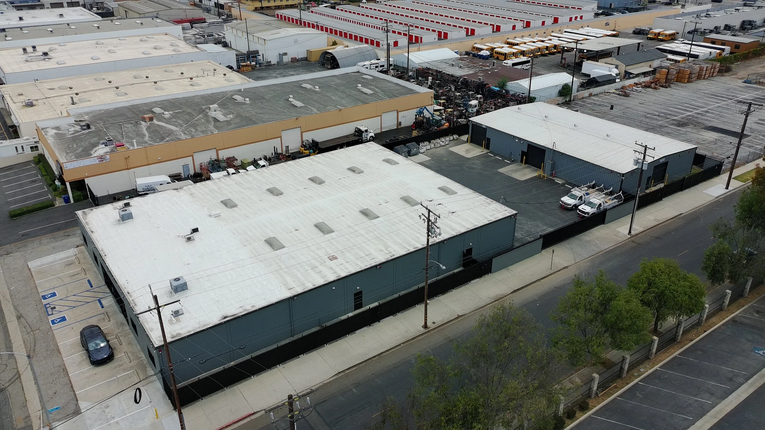 Aerial view of an industrial area with multiple flat-roofed warehouses, parking spaces, and a fenced lot containing various equipment and vehicles.