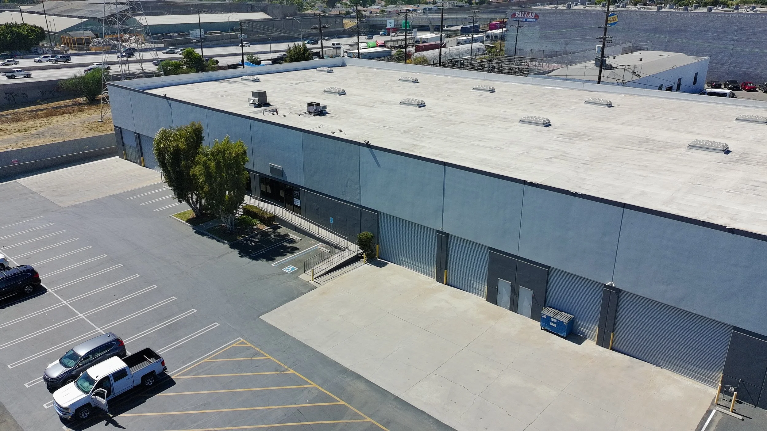 An aerial view of a large warehouse with a white flat roof, surrounded by a parking lot with some cars parked and vacant spaces. There are trees and a few smaller buildings nearby, and a busy highway with vehicles passing by in the background.