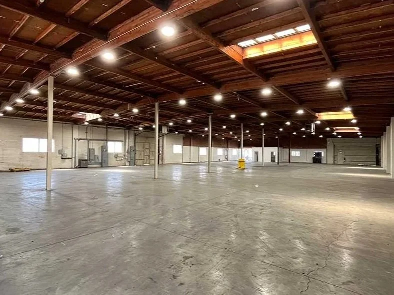 Empty commercial warehouse with a wooden ceiling and concrete flooring, illuminated by ceiling lights, featuring support poles and small windows along the walls.