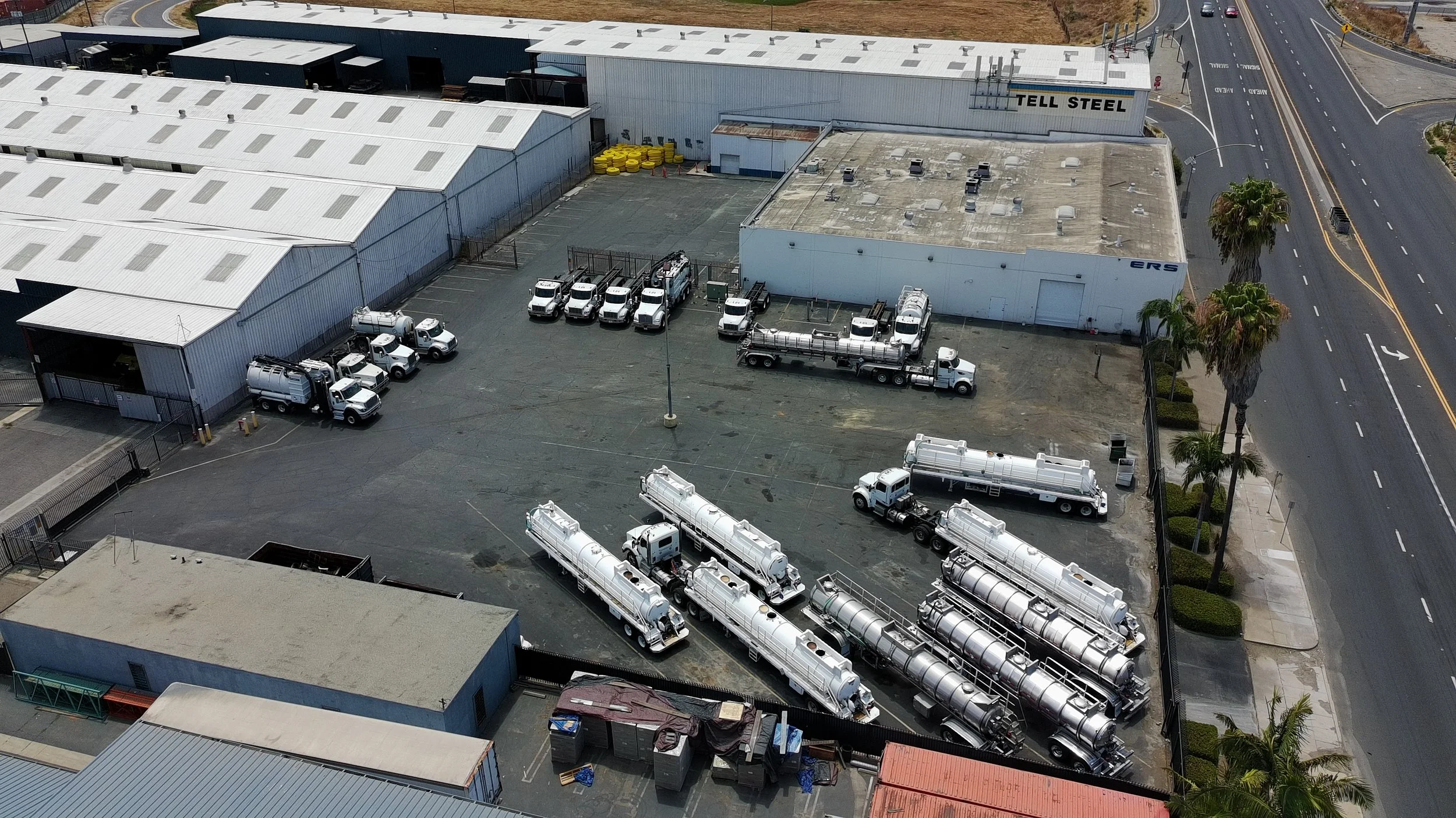An industrial facility with multiple large buildings, parked trucks and water tanks, surrounded by a fence, next to a road with palm trees.