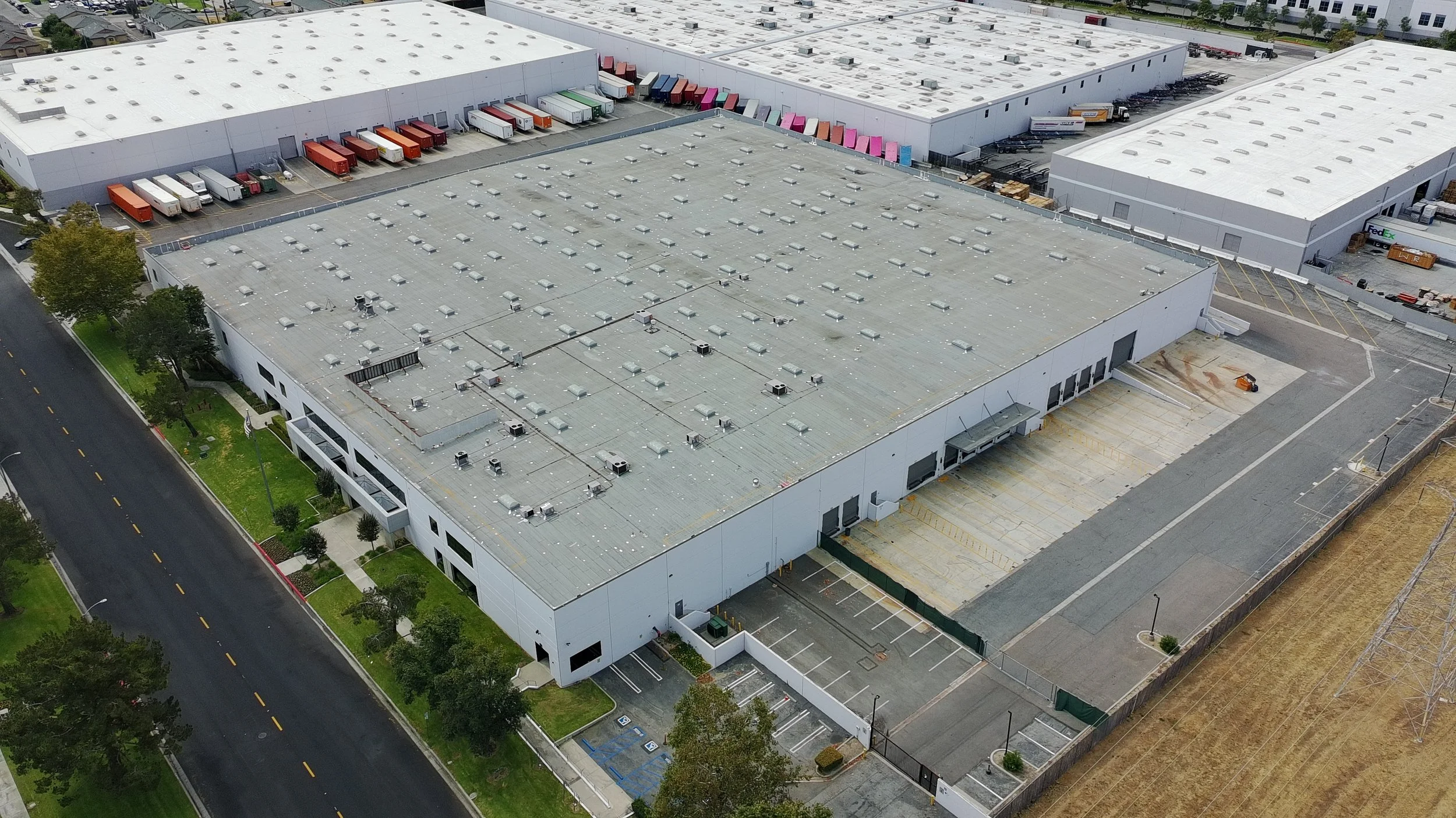 An aerial view of a large industrial building with a gray roof, surrounding parking lots, and several trucks parked at loading docks.