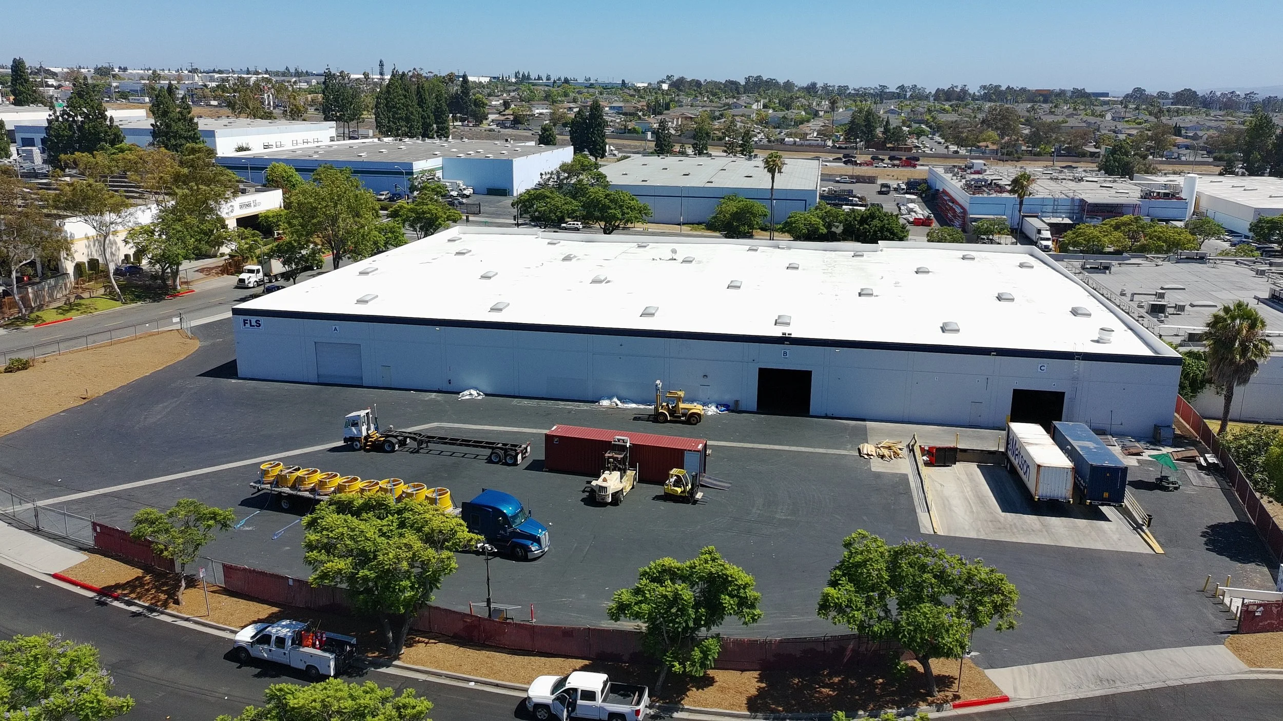 An aerial view of an industrial area with warehouses, trees, parking lots, trucks, and construction equipment under a clear blue sky.