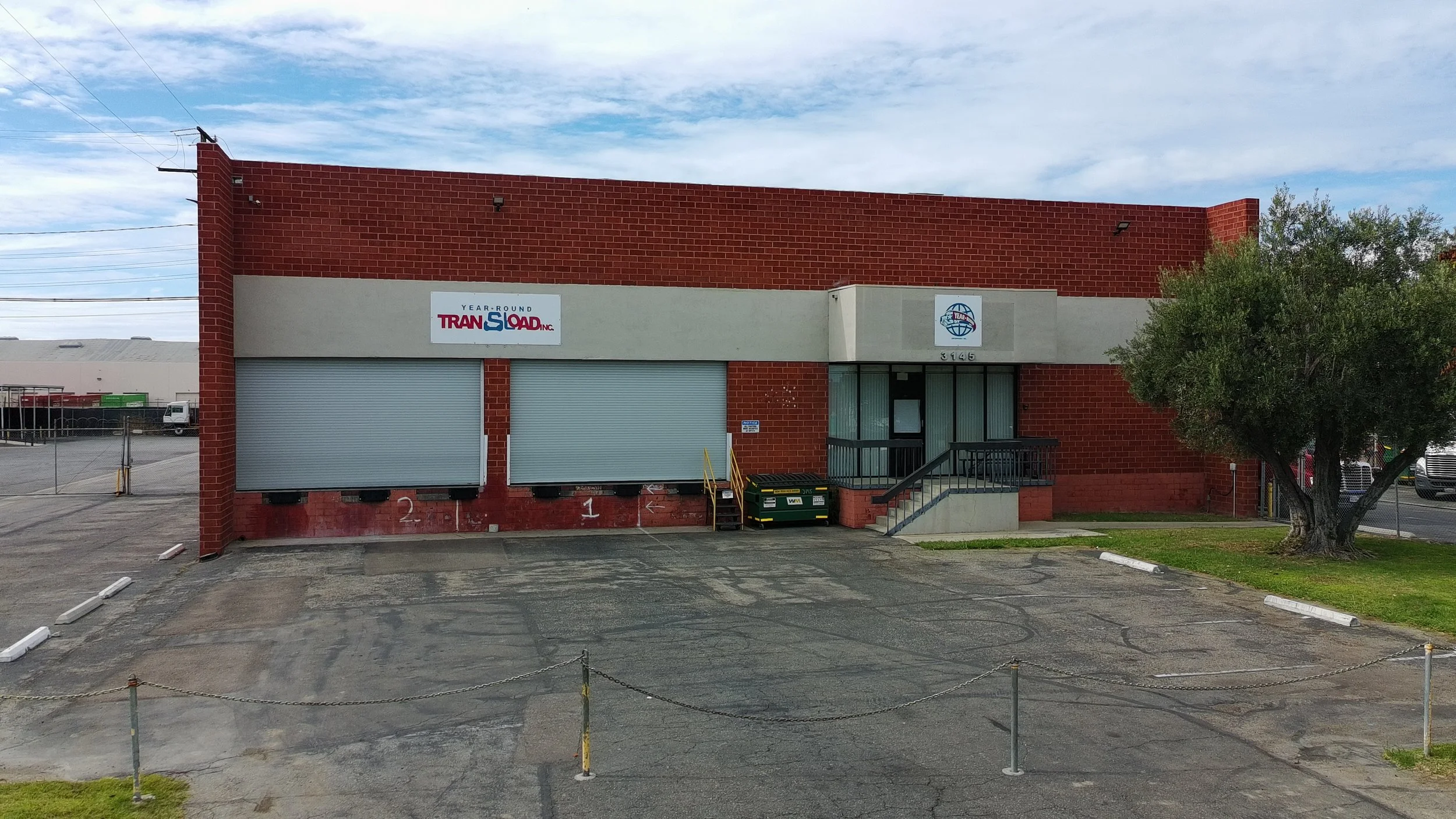 A two-story commercial building with a brick upper floor and a white lower section. There are two closed metal roll-up doors on the lower level, numbered 1 and 2. To the right, the entrance features a glass door with a small staircase and black raili