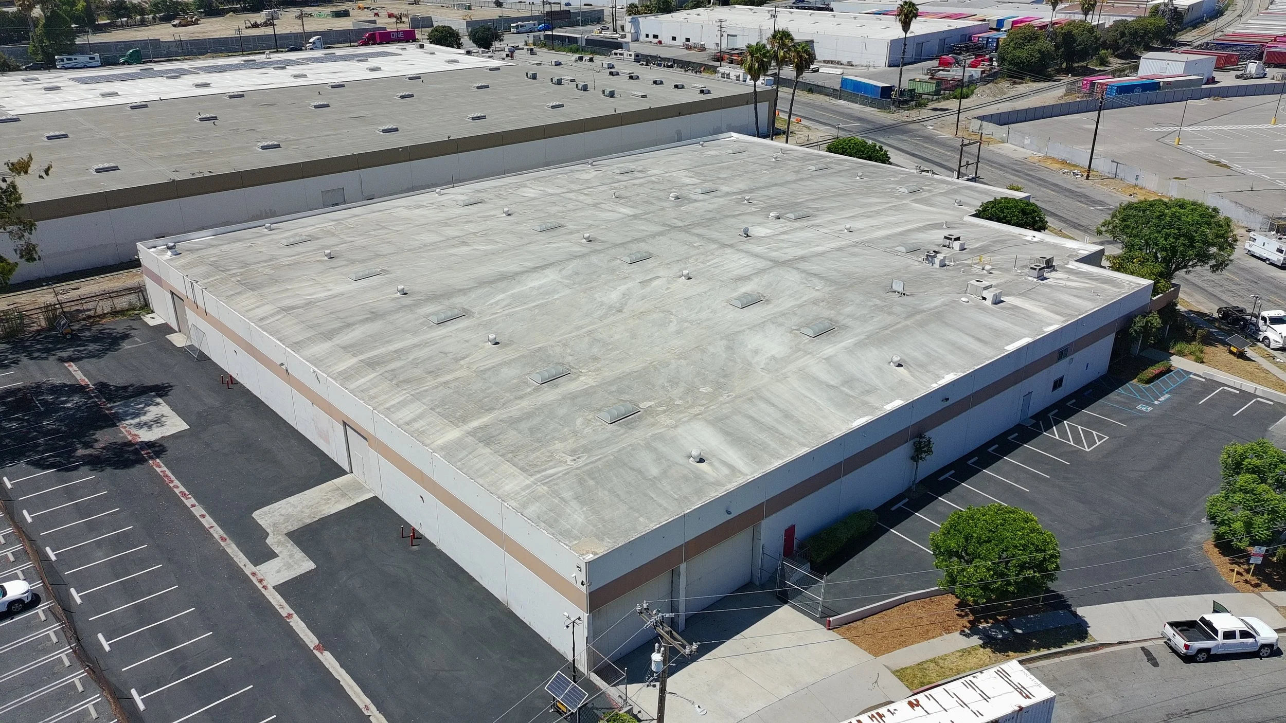 Aerial view of a large commercial building with a flat, gray roof, parking lot, and surrounding trees and streets.