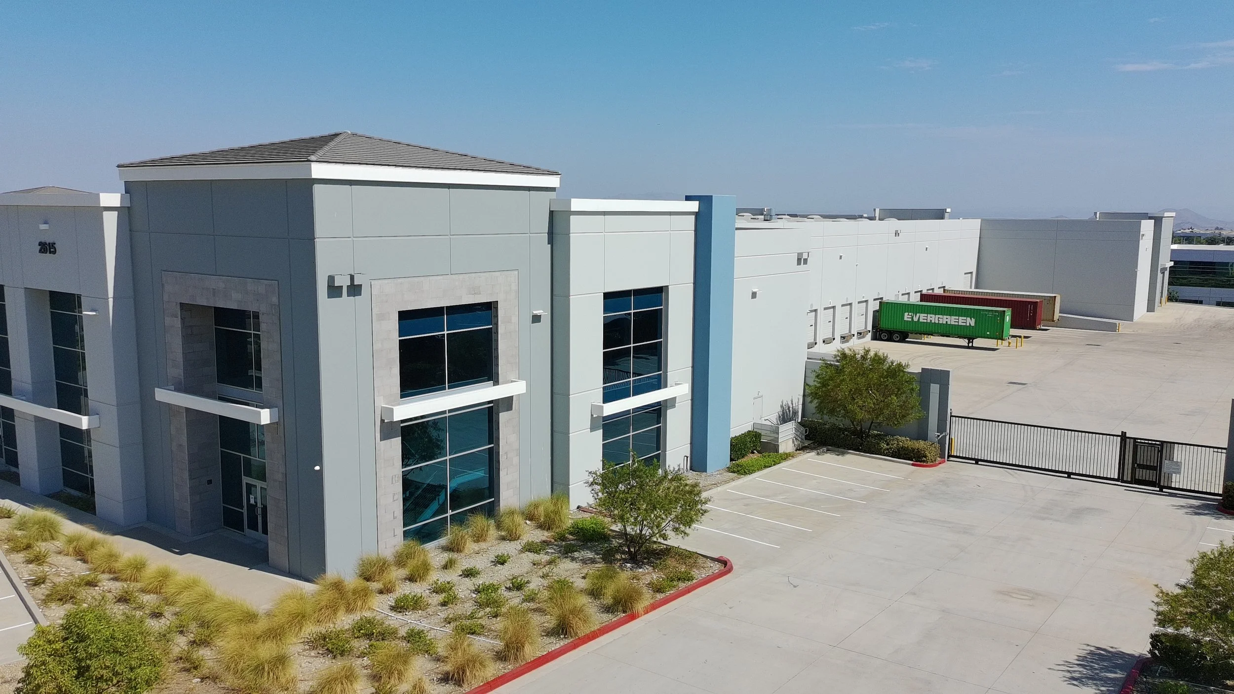 Modern commercial building with large windows and landscaping in the foreground. Parking area with a green and a red shipping container nearby.