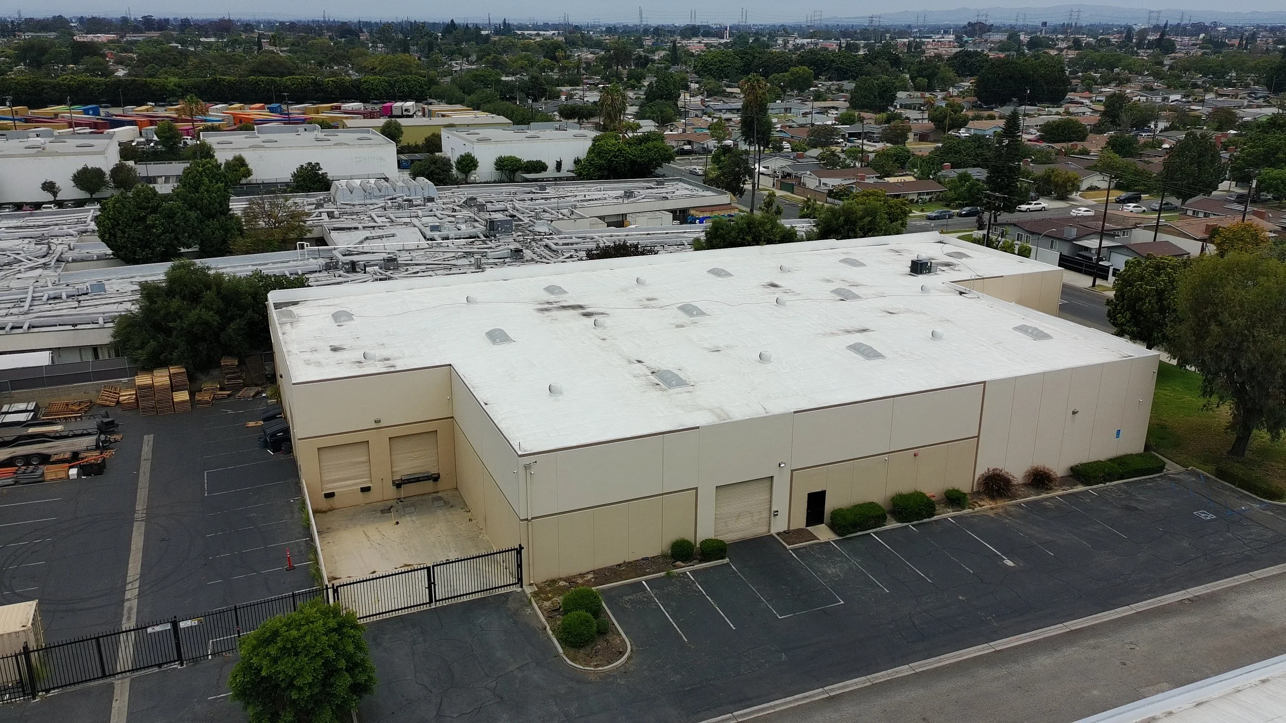 Aerial view of a large commercial building with a white rooftop, surrounded by parking lots, trees, and an urban neighborhood with residential houses in the background.