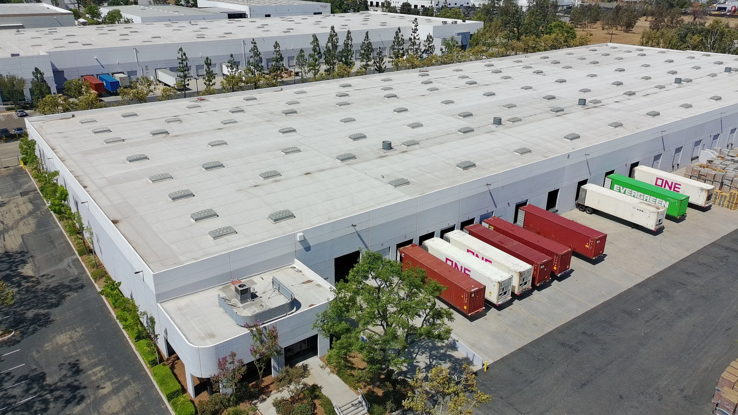 Aerial view of a large warehouse building with a white roof, multiple vents, and several trucks parked at loading docks with their doors open.