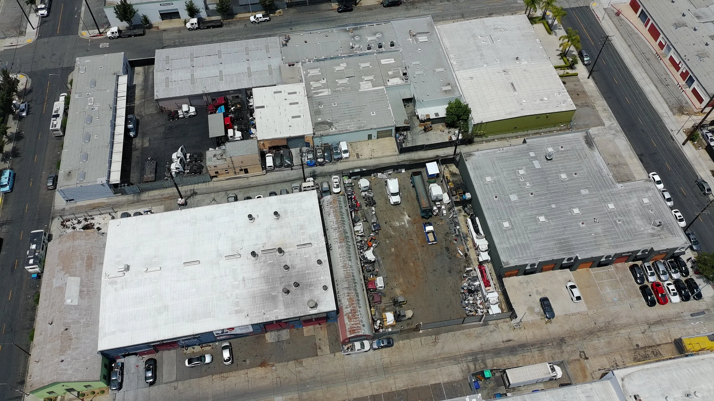 Aerial view of commercial buildings with flat roofs, parking lots filled with cars, and a street with vehicles and palm trees on the sidewalk.