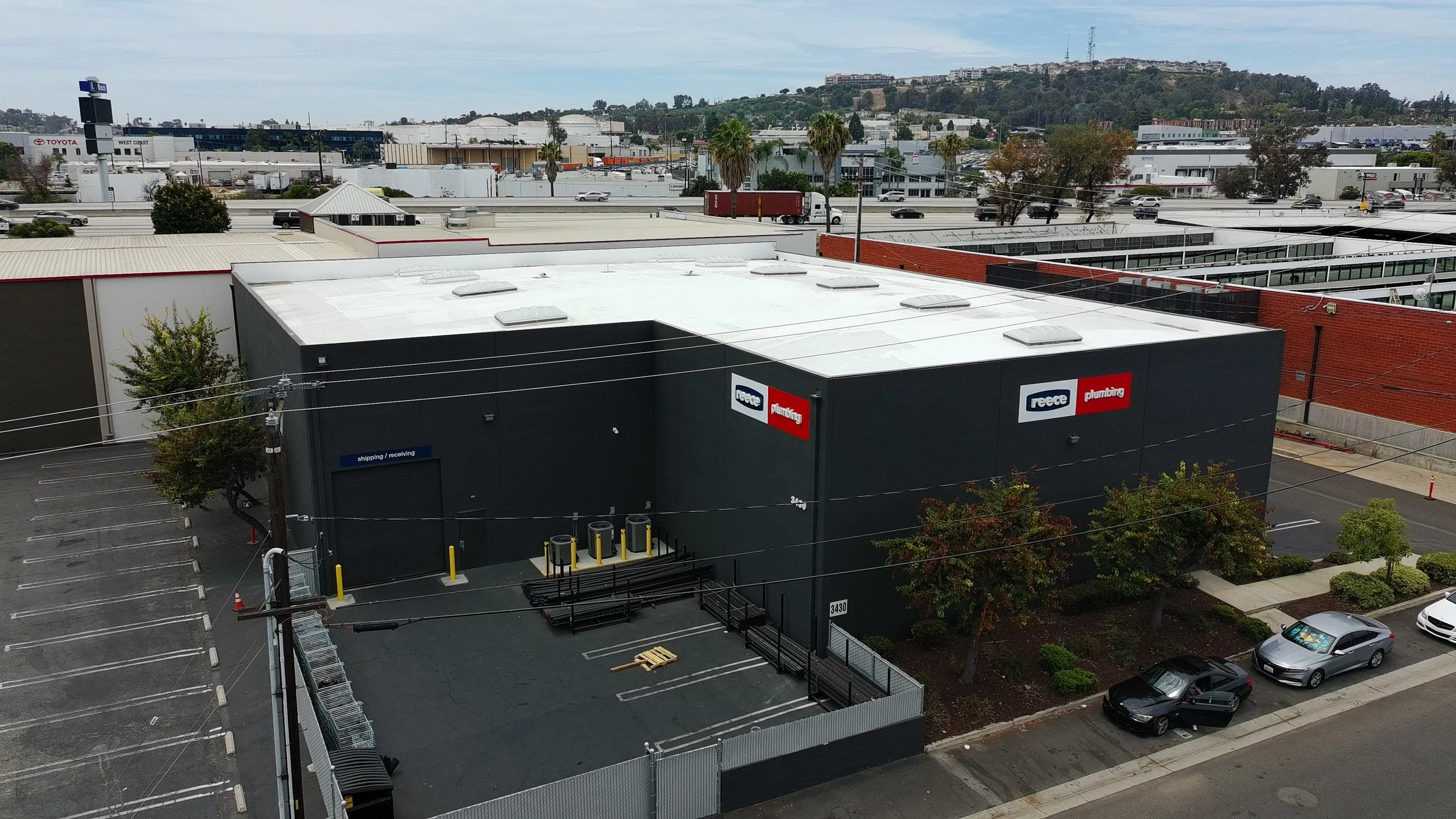 An industrial building with signs that read 'Reece Plumbing' and 'shipping/receiving' in an urban area with parking lot, trees, and a distant view of hills with buildings and communication towers.