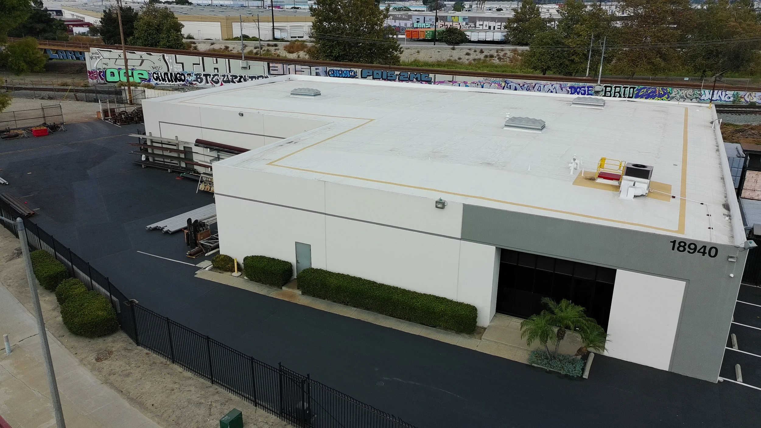 Aerial view of a large white commercial building with a flat roof, surrounded by a parking lot and a black fence, graffiti-covered train tracks behind the building, and some greenery.