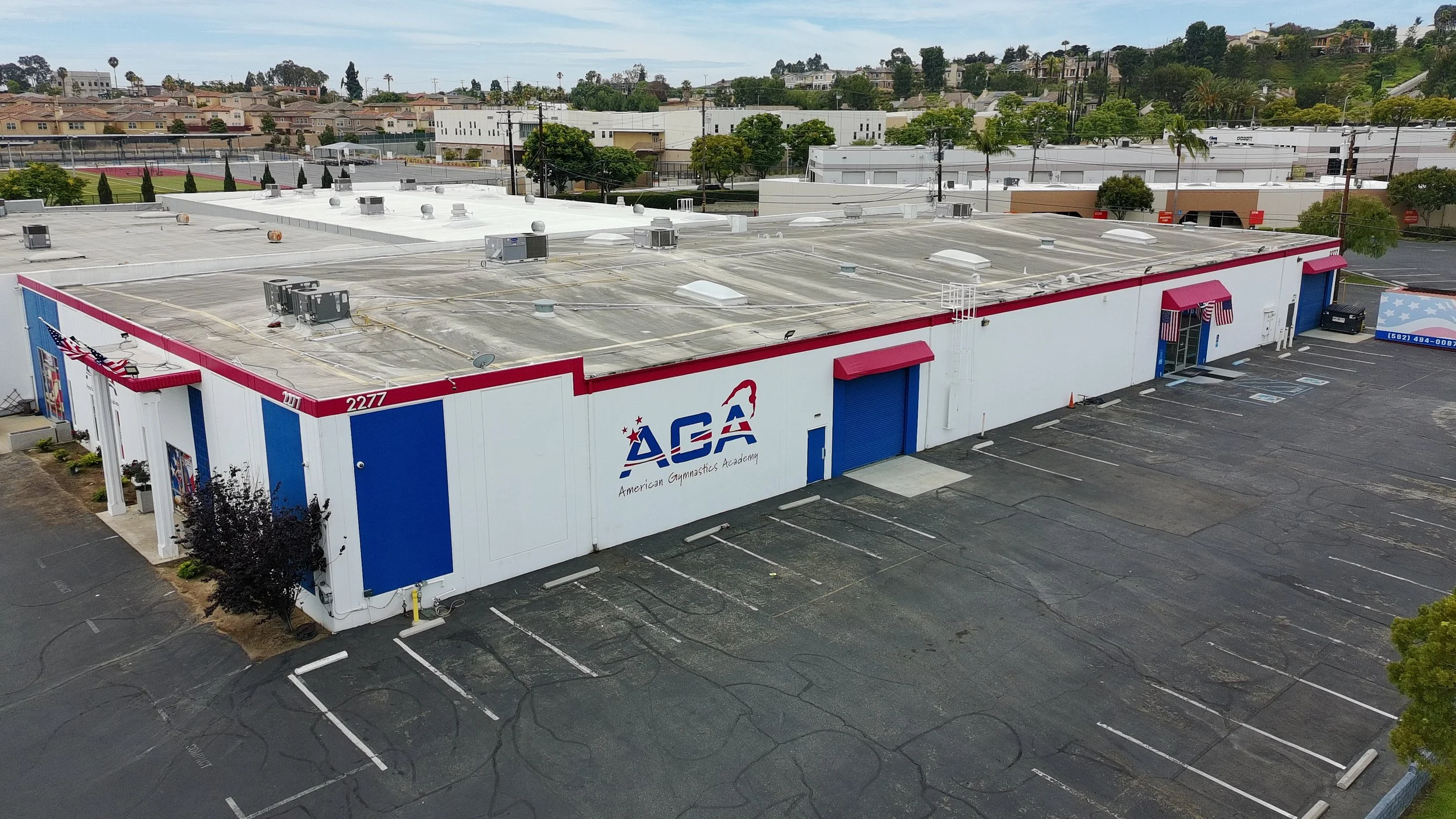 Aerial view of a large building with the logo 'ACA' for American Gymnastics Academy, surrounded by empty parking lot, and a hillside with residential houses in the background.