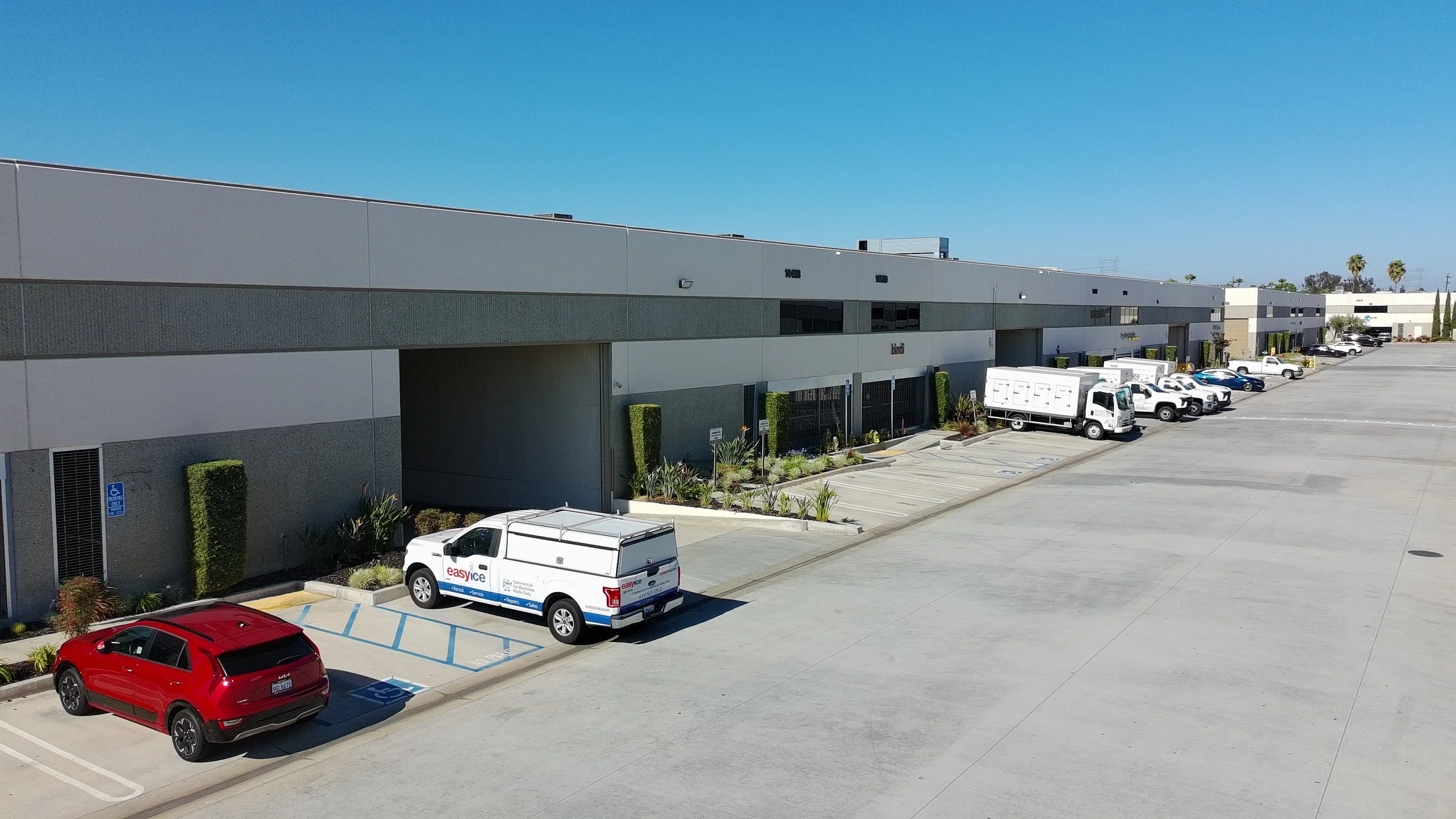 A large commercial building with a parking lot in front, showing parked vehicles including a white van and a red car, under a clear blue sky.