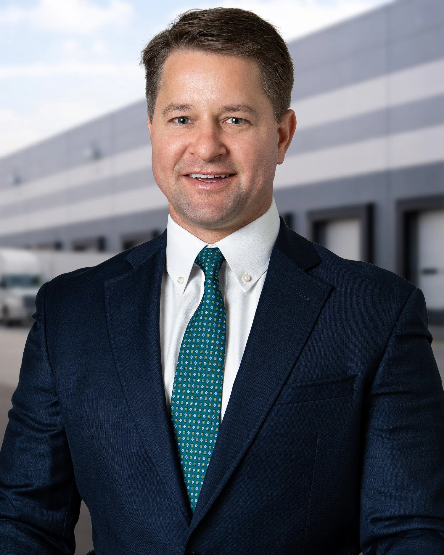 Professional headshot of a man wearing a navy suit, white shirt, and patterned blue tie, standing outdoors with a modern building in the background.