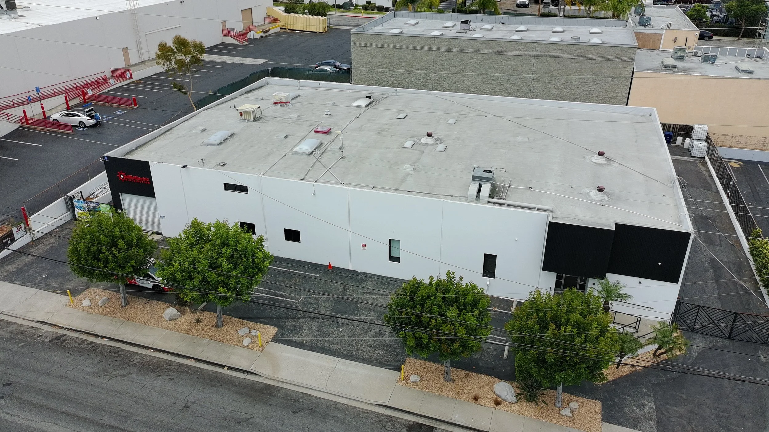 Aerial view of a commercial building with a white roof, parking lot with parked cars, trees, and neighboring buildings.