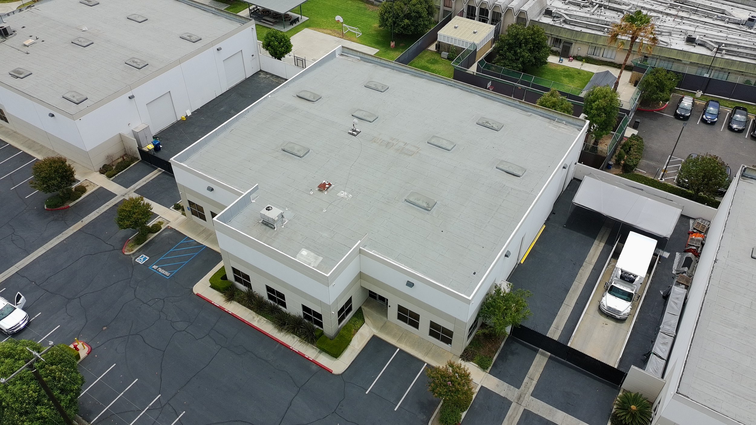 An aerial view of a commercial building with a flat gray roof, parking lot, and surrounding greenery. Several cars are parked, and there are trees and a small sports court nearby.