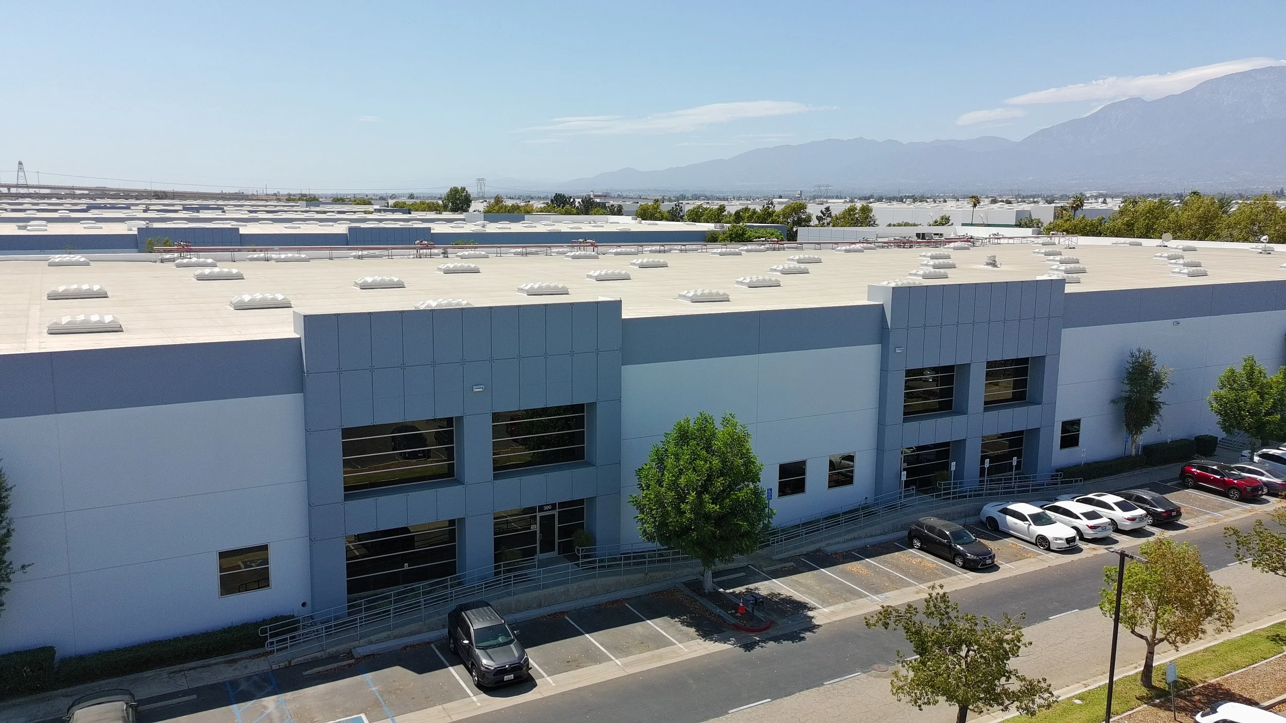 View of a commercial building with a flat roof, parking lot, and cars. Distant mountains and partly cloudy sky in the background.