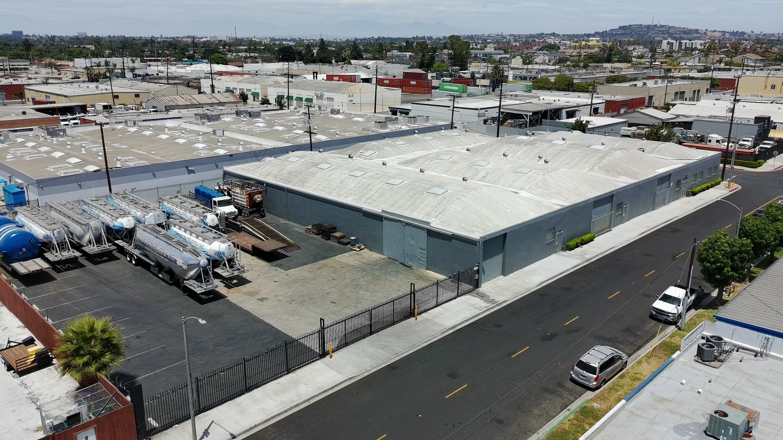 Aerial view of an industrial area with large warehouse buildings, parked cars, semi-trucks, and water tanks on rooftops, with residential and commercial buildings in the background under a partly cloudy sky.