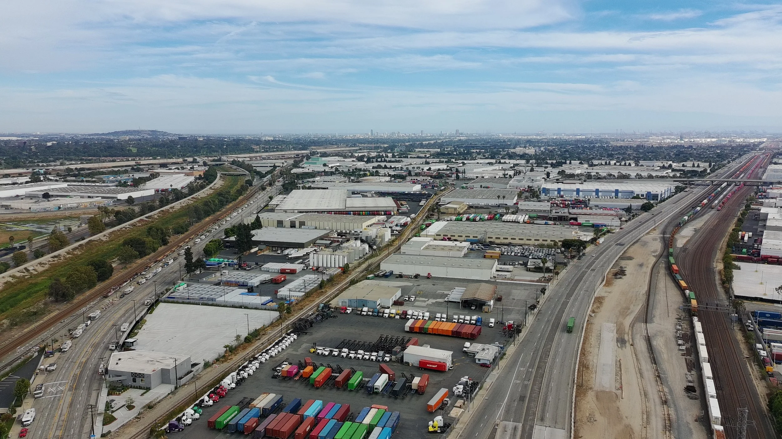 Aerial view of a large industrial and warehouse area with numerous shipping containers, railways, roads, and buildings under a partly cloudy sky.