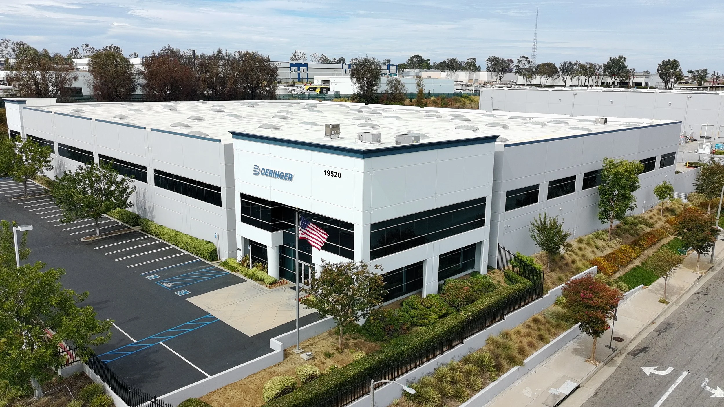 A large white commercial building with the company name 'Deringer' and address '19520' on the front. It has a parking lot with marked spaces, trees, and well-maintained landscaping, with some trees displaying fall colors.