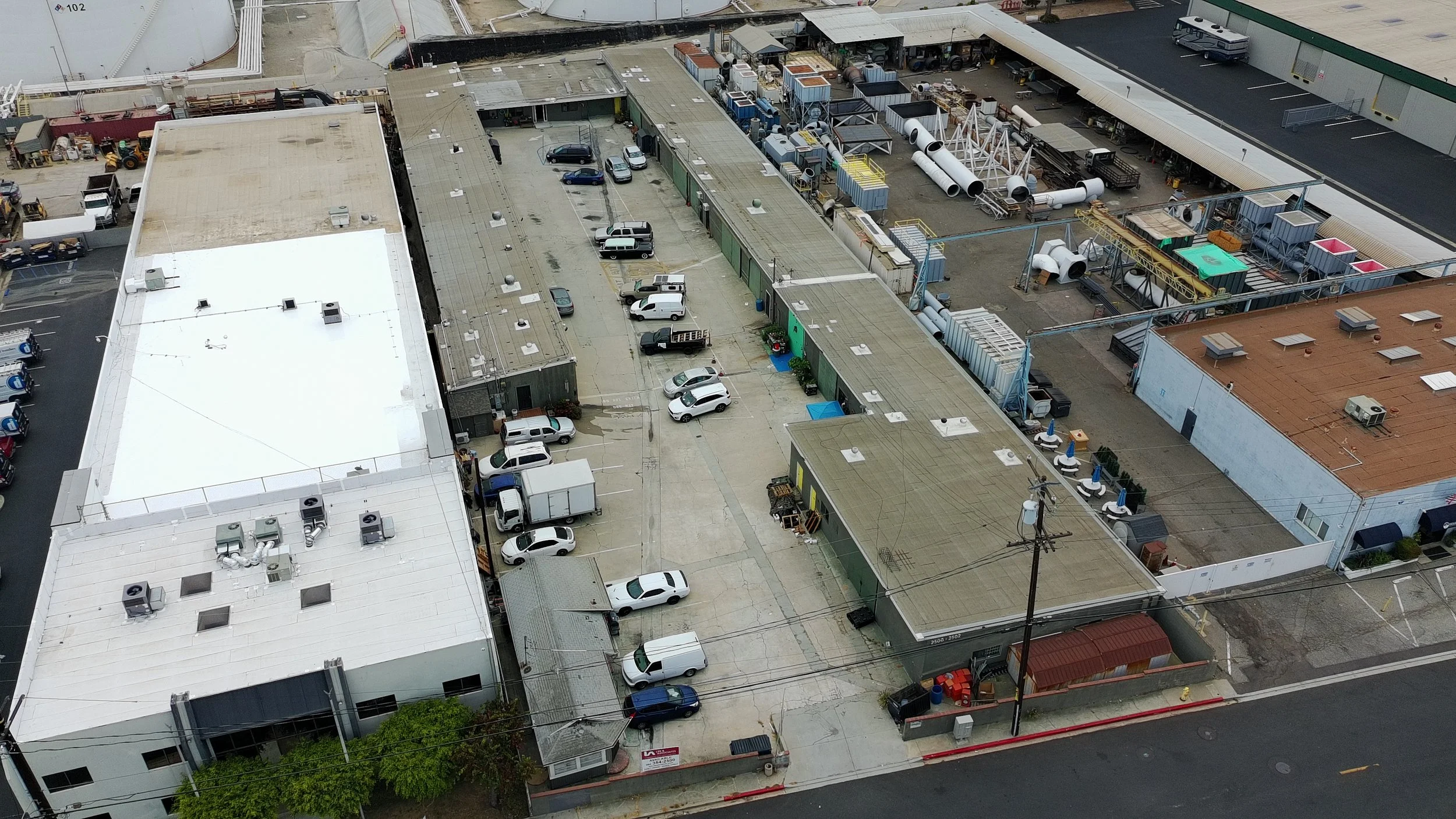 Aerial view of an industrial area with multiple buildings, parking lot, and outdoor storage of pipes and equipment.