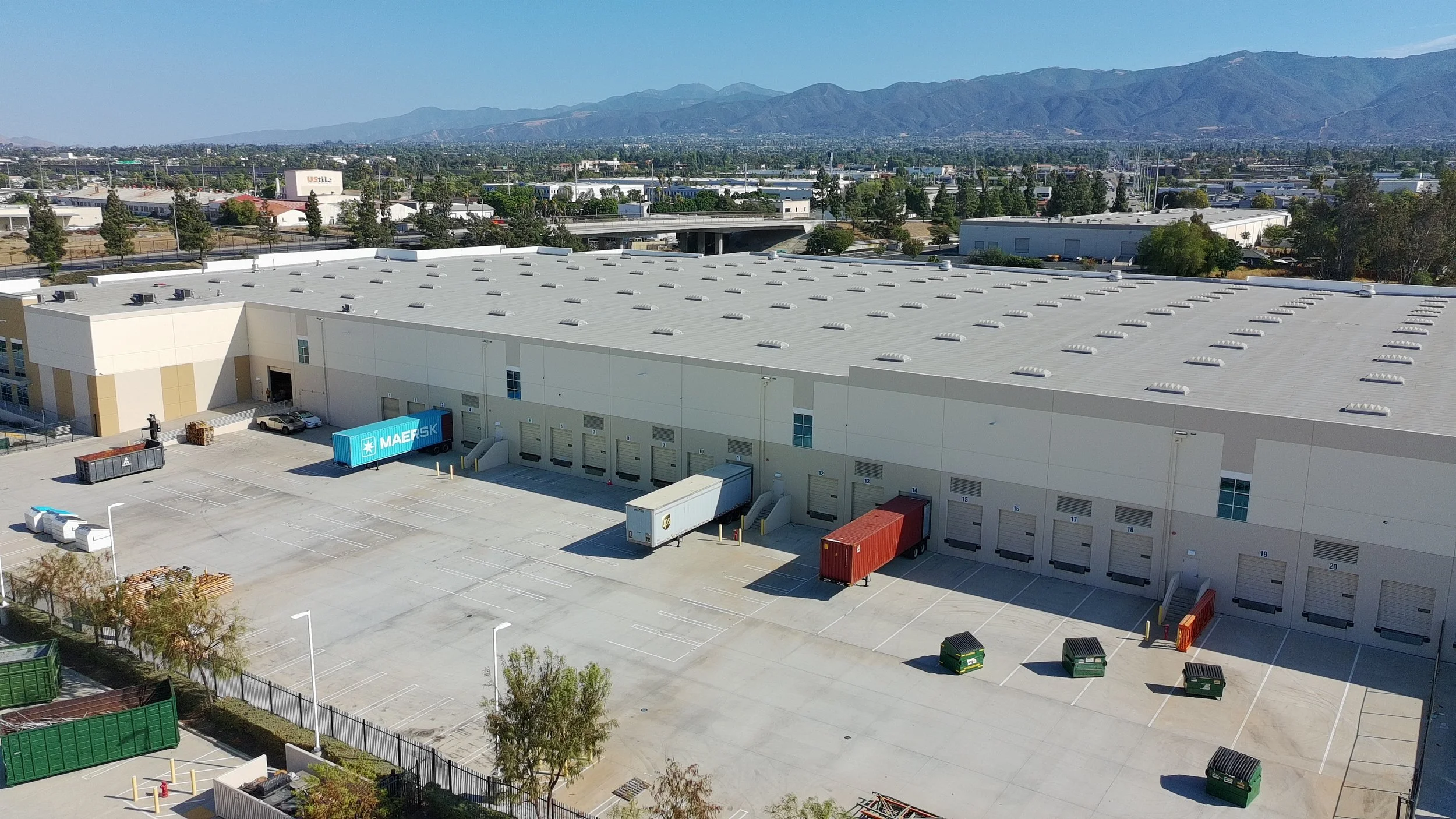 A large warehouse with loading docks and trucks, set against a backdrop of mountains and a partly cloudy sky.