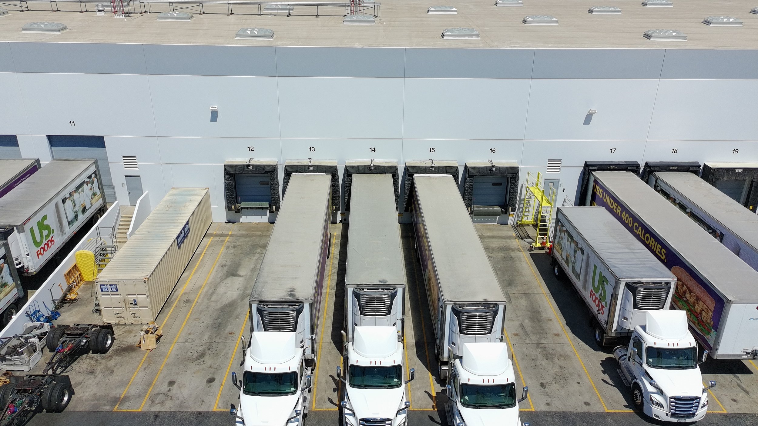 Aerial view of the loading dock area with several trucks parked in designated spaces, some trucks are labeled with advertising for US Foods, and loading doors numbered from 11 to 19 on a large warehouse building.