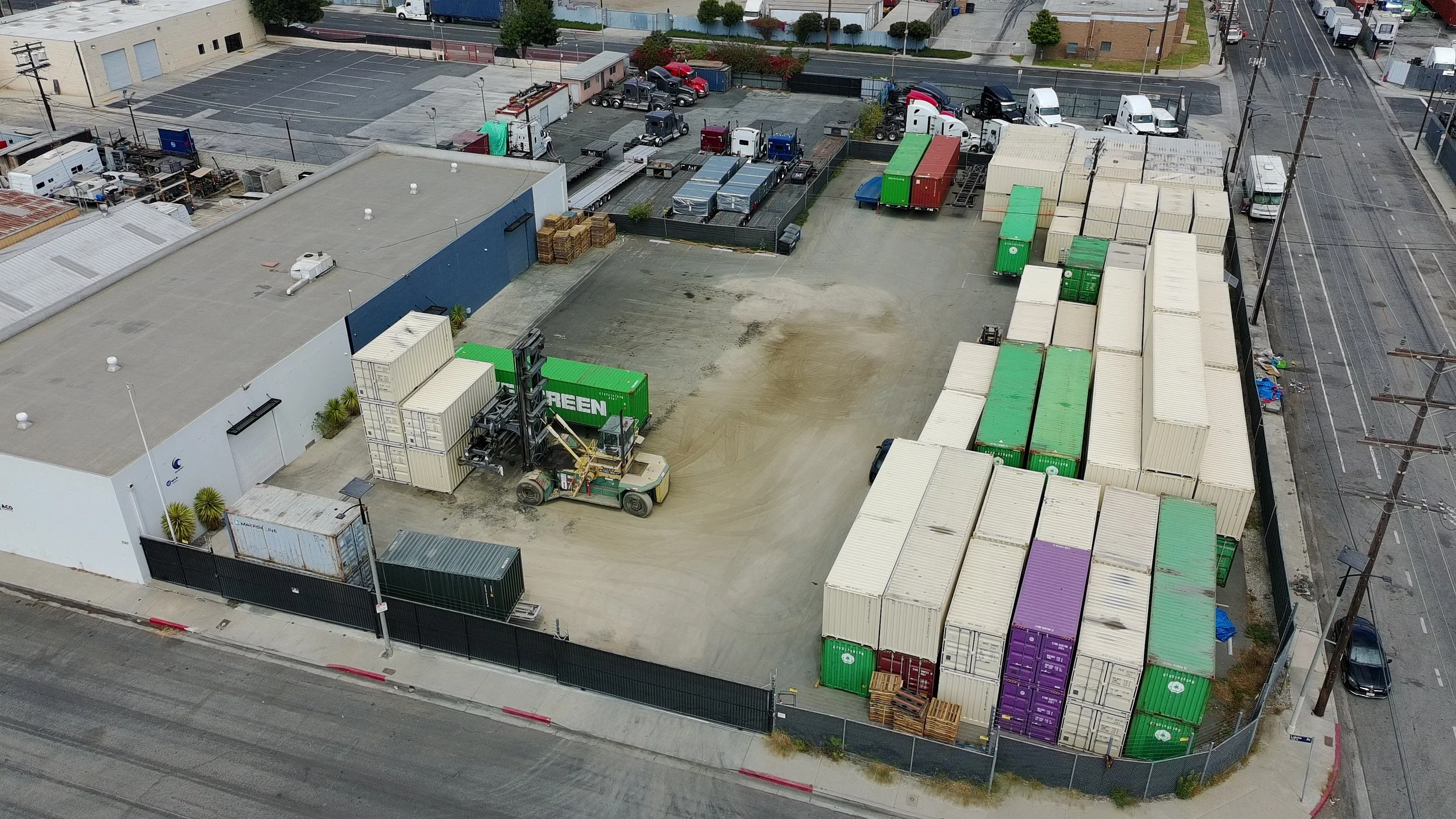 Aerial view of a parking lot and storage area with numerous shipping containers, trucks, and industrial equipment, enclosed by fences and located next to roads and buildings.