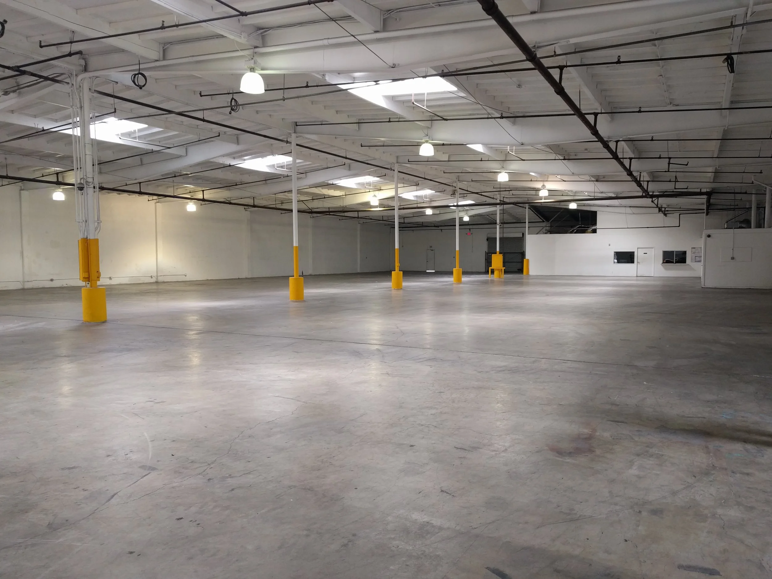 Empty indoor parking garage with yellow safety bollards, white ceiling with overhead lighting, and exposed pipes and electrical conduits.