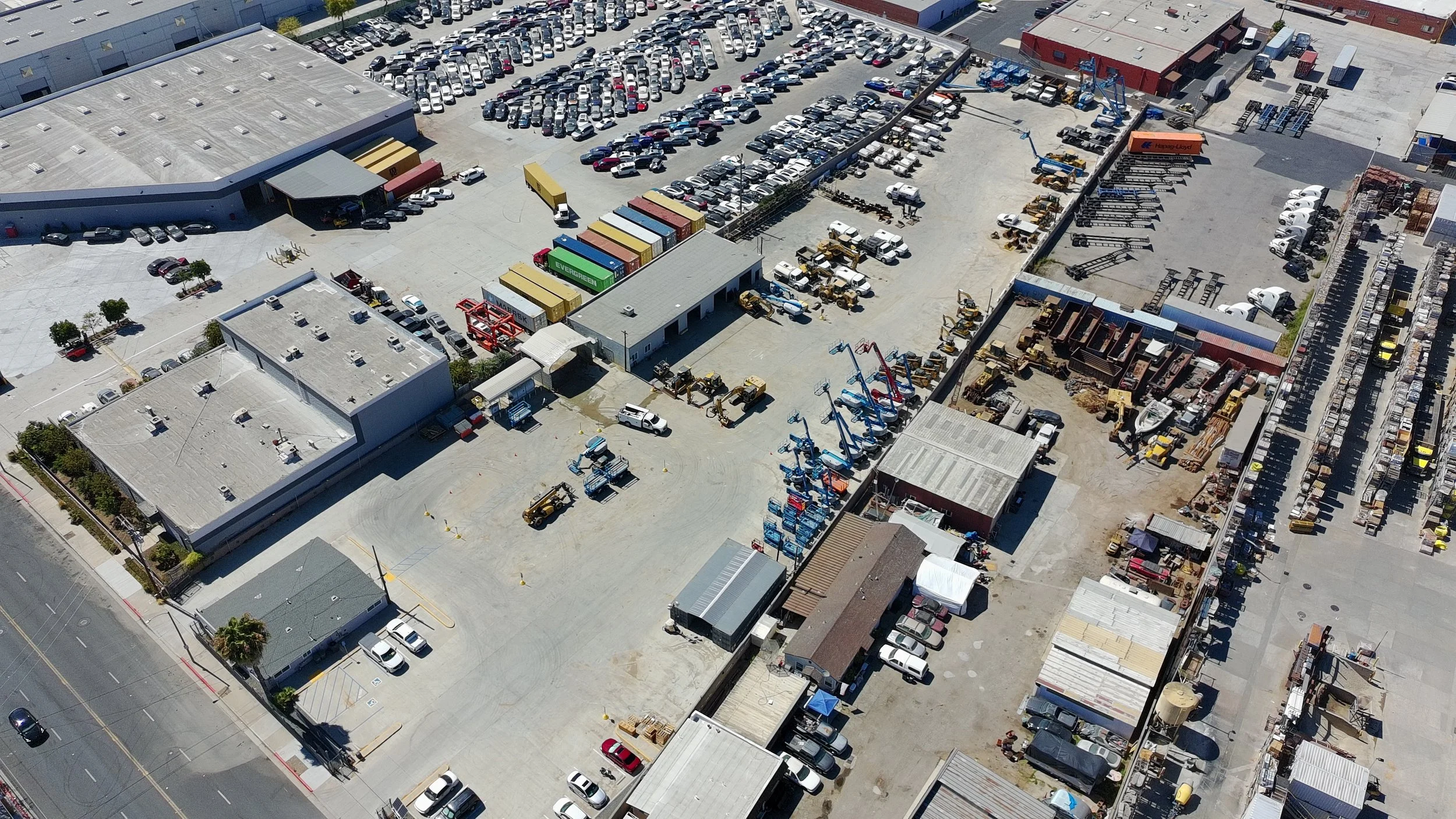 An aerial view of a commercial yard with parked cars, trucks, and construction equipment, including cranes and trailers, with surrounding buildings and streets.