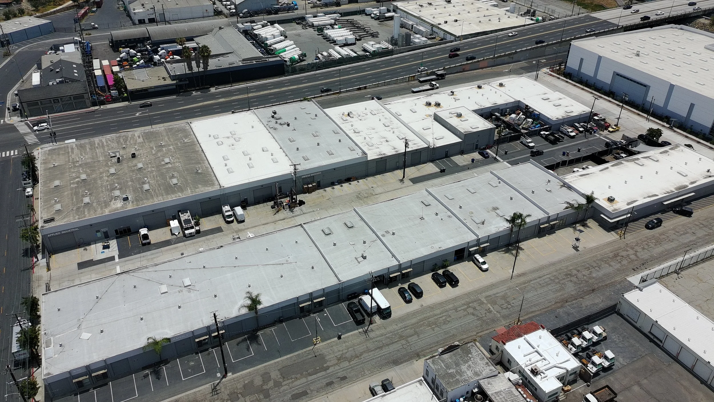 Aerial view of a commercial area with multiple flat rooftops, parking lots, and a busy street with cars. The buildings include warehouses and retail stores.