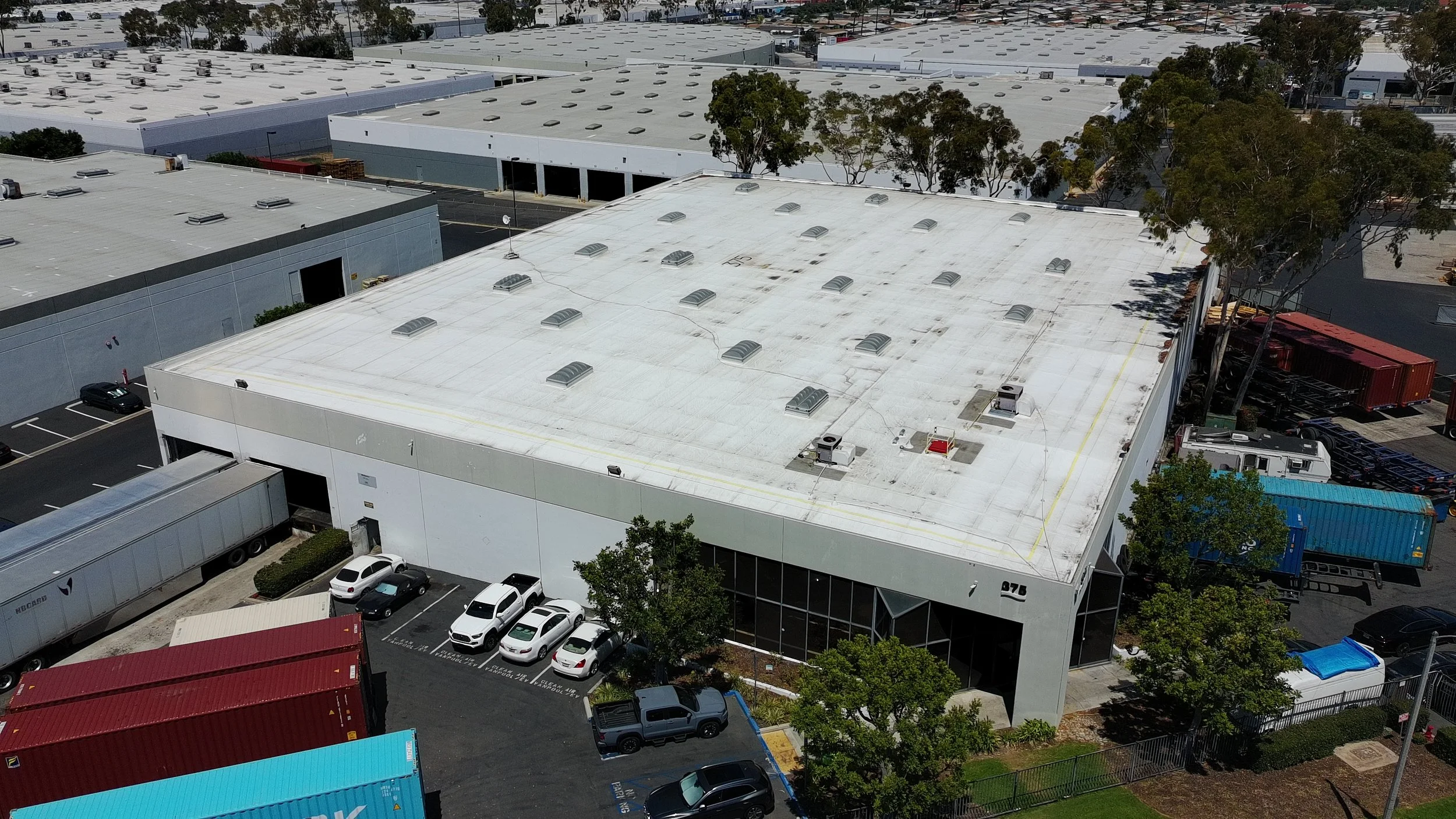 An aerial view of large commercial warehouse buildings with parking lots and trees surrounding the area, taken during daytime.