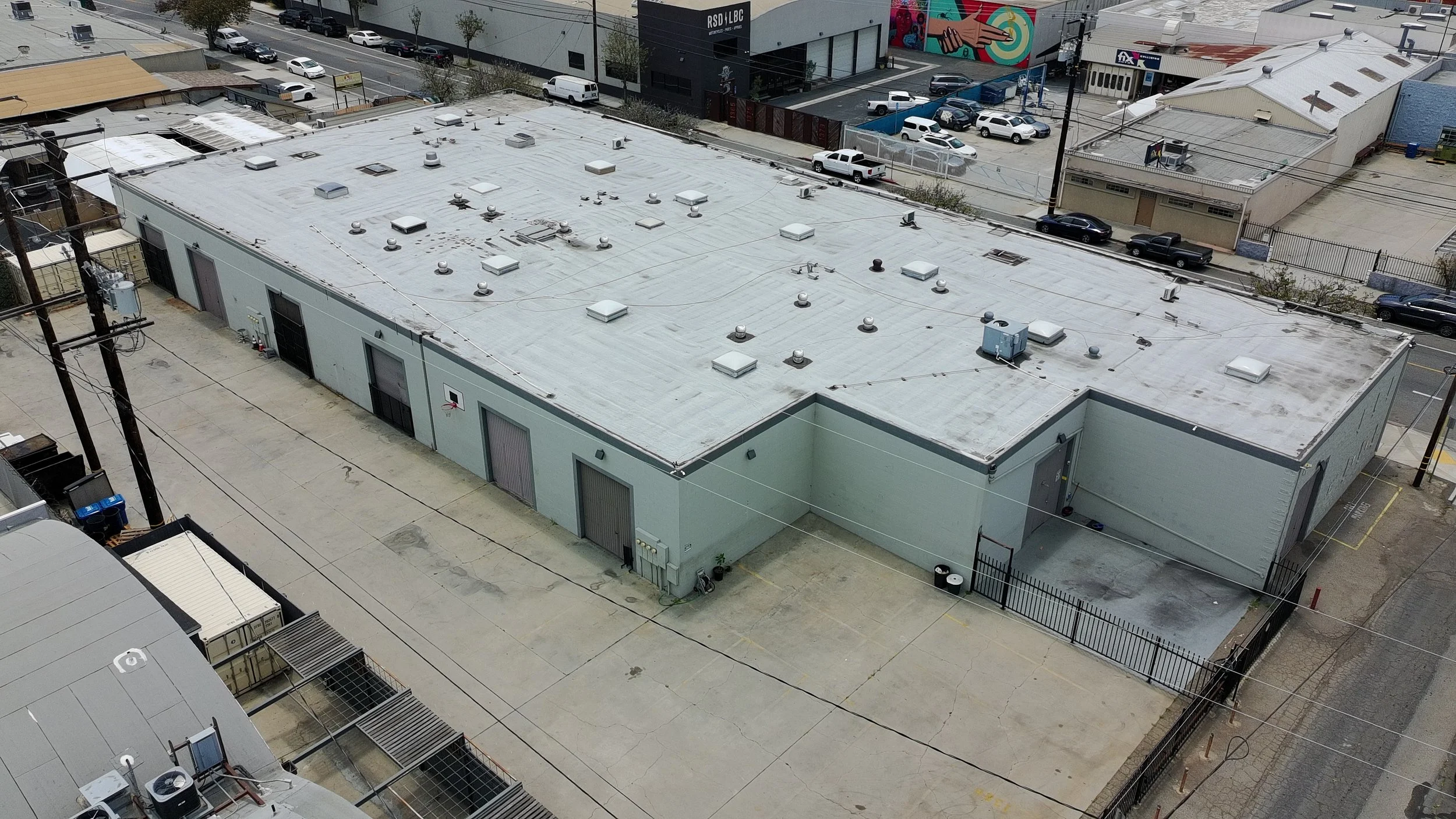 A flat rooftop of a commercial building with multiple vents, skylights, and HVAC units, surrounded by a paved area and nearby streets with parked cars.