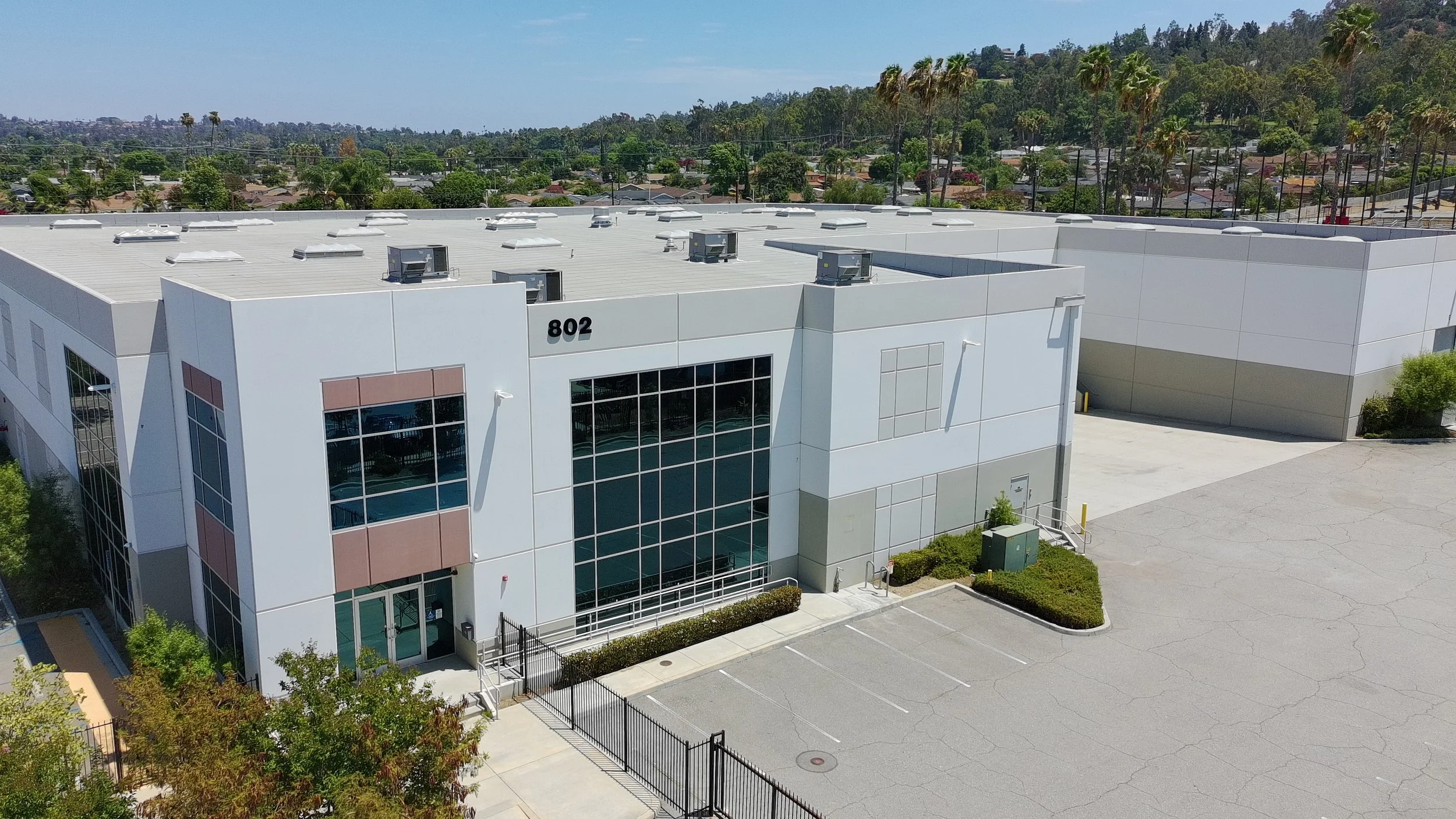 An aerial view of a modern commercial building with a flat roof and large glass windows, surrounded by a parking lot and some trees, with a residential area and forested hills in the background.
