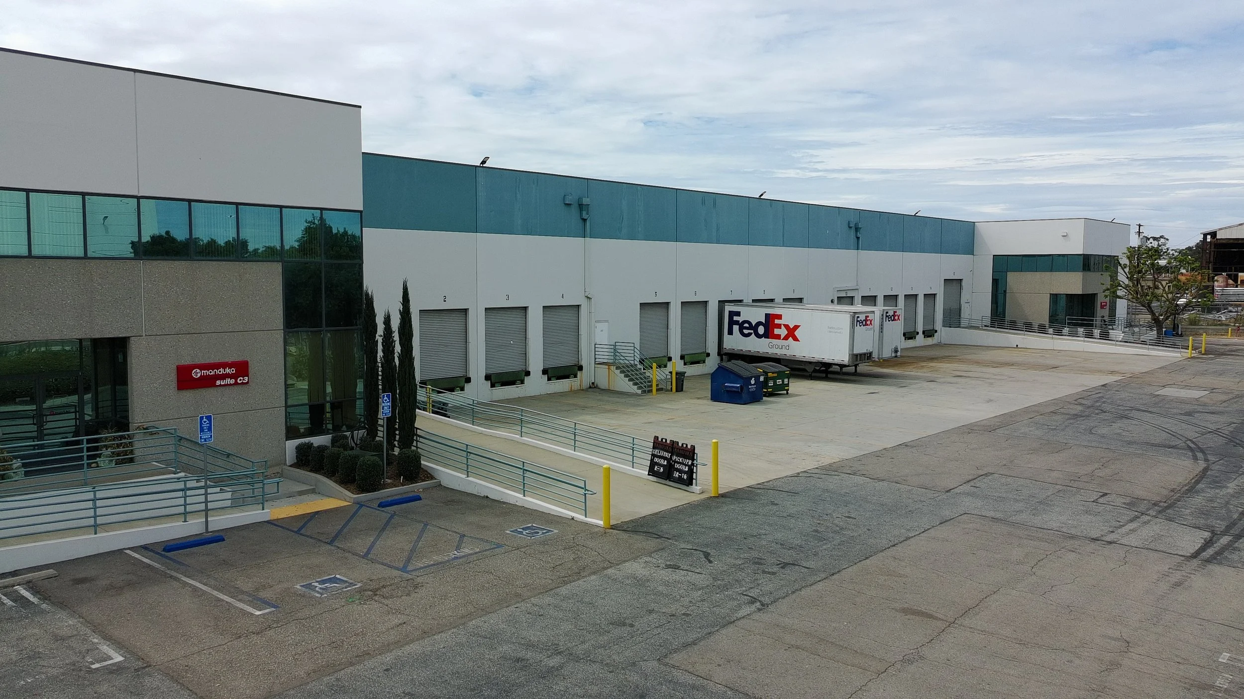 A large warehouse or distribution center with loading docks, a FedEx truck parked outside, and a few parking spaces in the foreground.