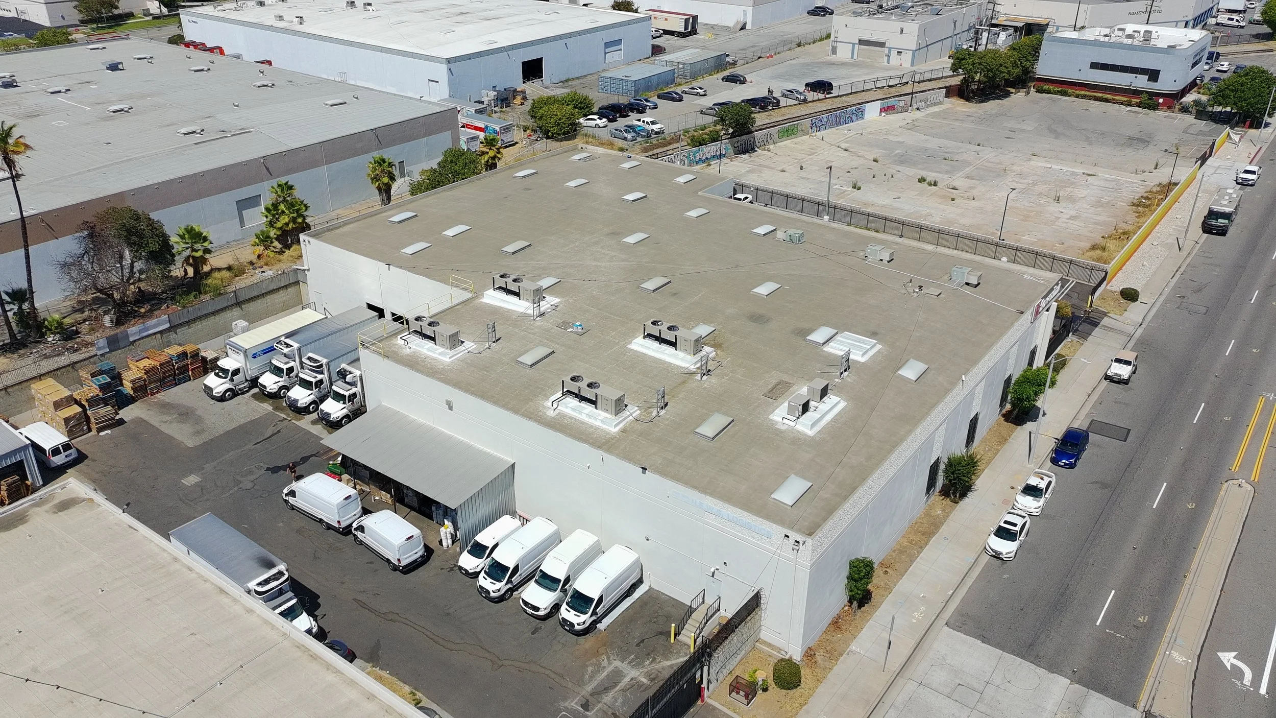 Aerial view of a commercial building with a flat roof, surrounded by parked vans, cars, and trucks, with an empty parking lot and graffiti on the fence in the background.